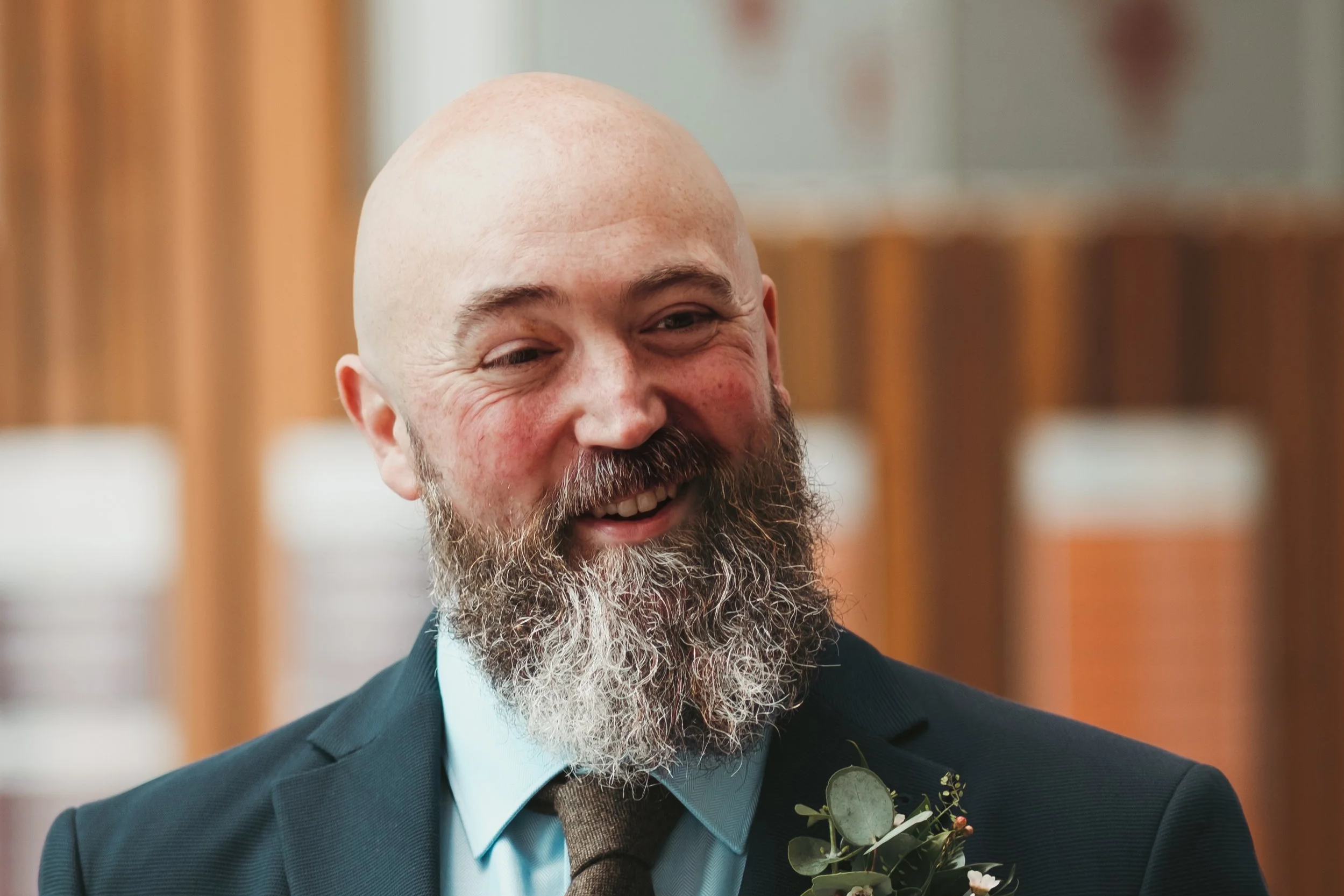 A smiling bald man with a beard dressed in a dark suit, light blue shirt, and brown tie, wearing a boutonniere with greenery and small flowers, standing indoors with wooden paneling and cubical storage units in the background.
