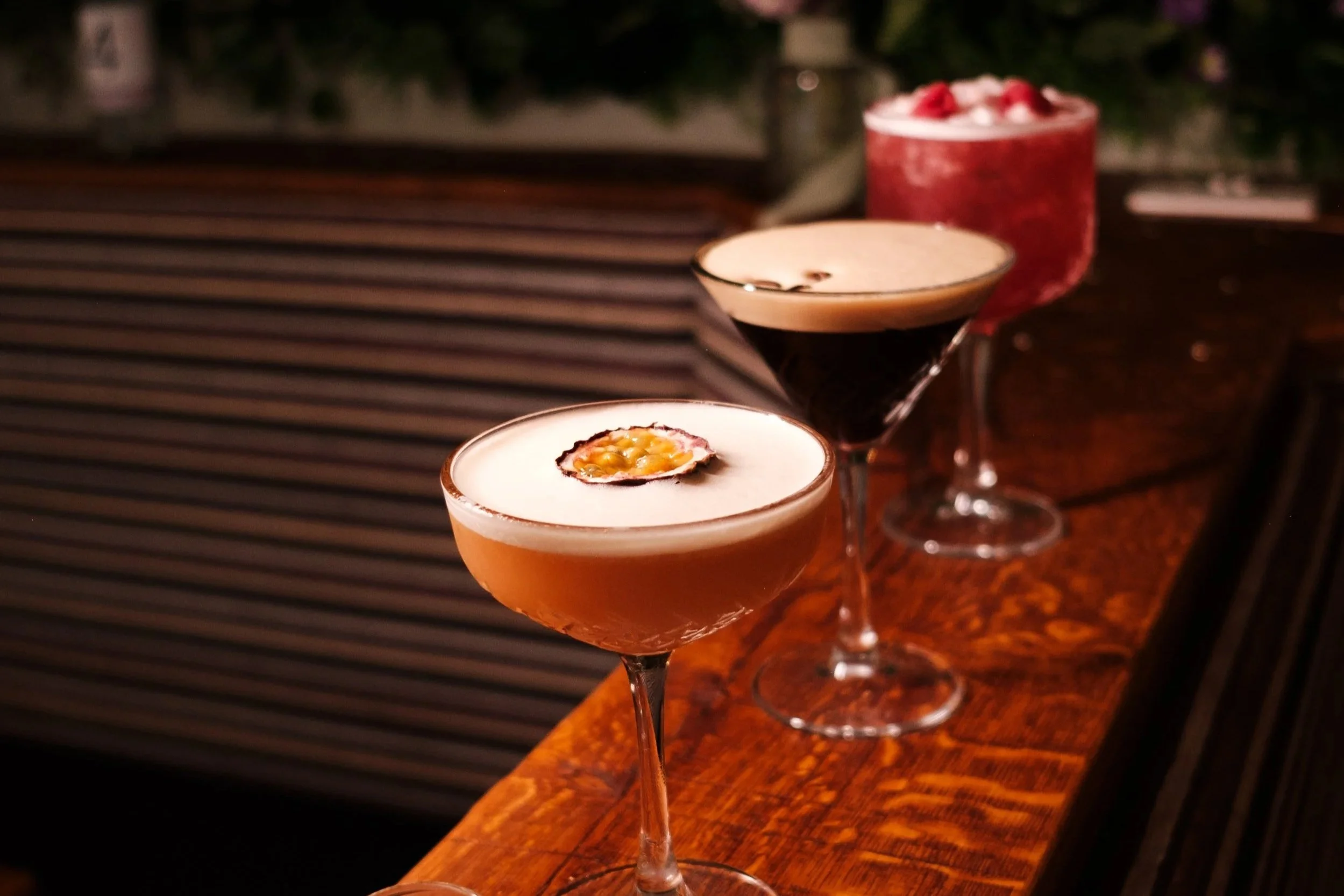 Three cocktails on a wooden bar counter with a floral background.