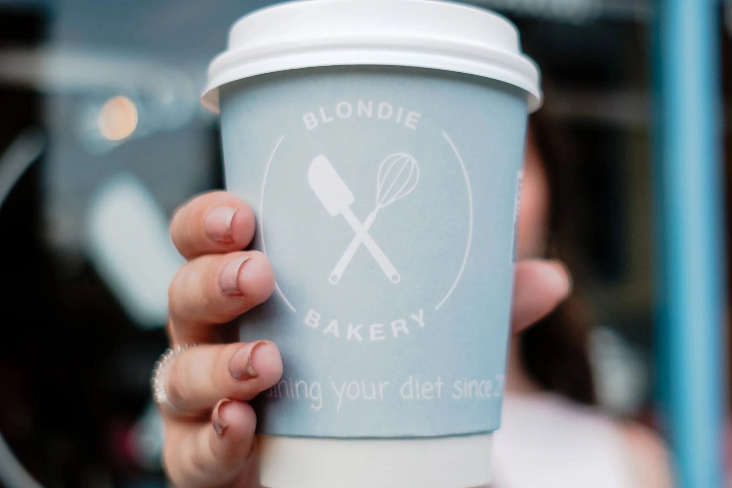 Person holding a blue paper coffee cup with a white lid, featuring a bakery logo with a spatula and whisk crossed, outside a bakery storefront.
