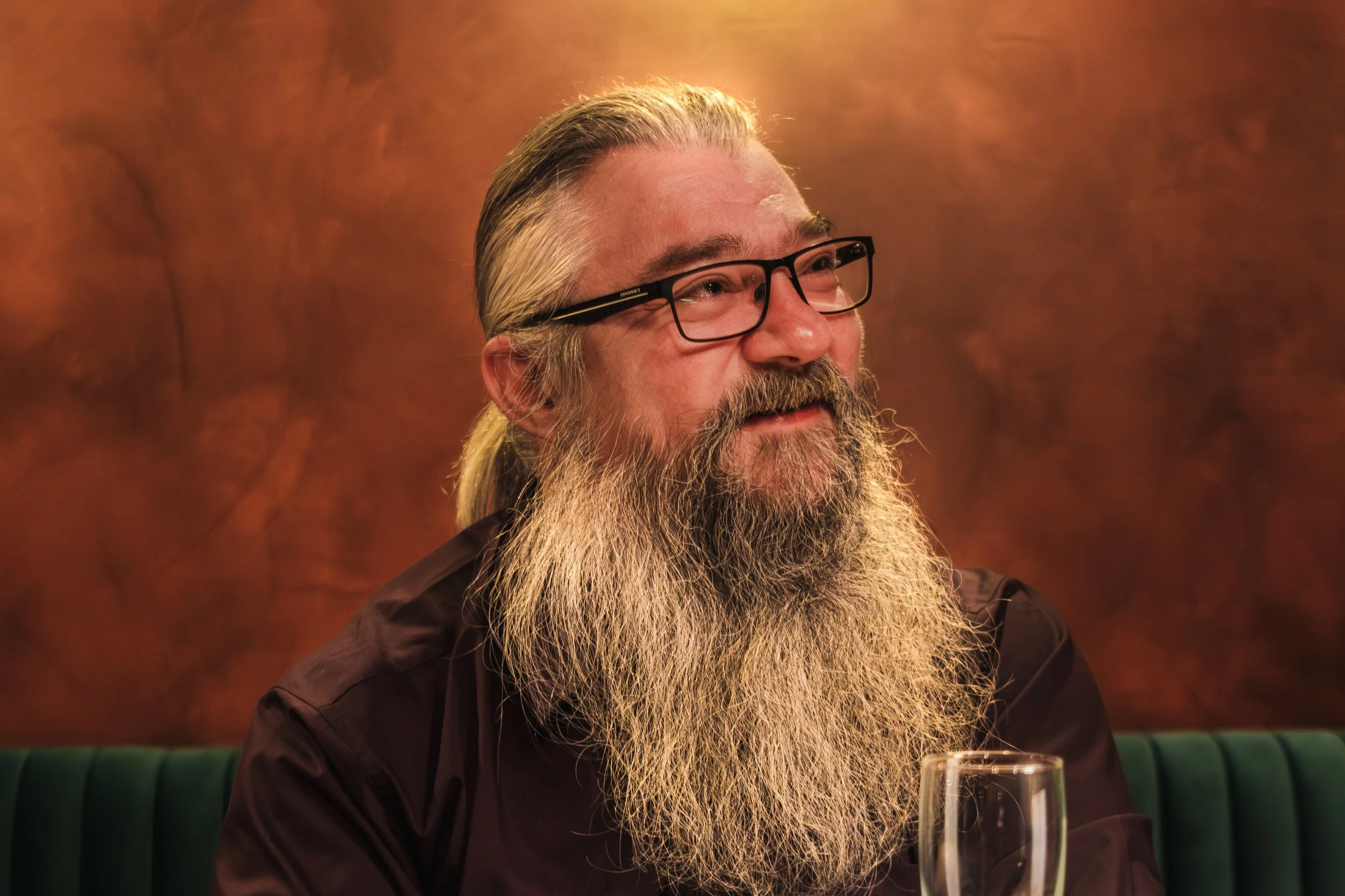 A man with a long gray beard, glasses, and tattoos on his arms sitting at a wooden table with a glass of champagne in front of him. The background has warm-colored walls and a clock hanging on the wall.