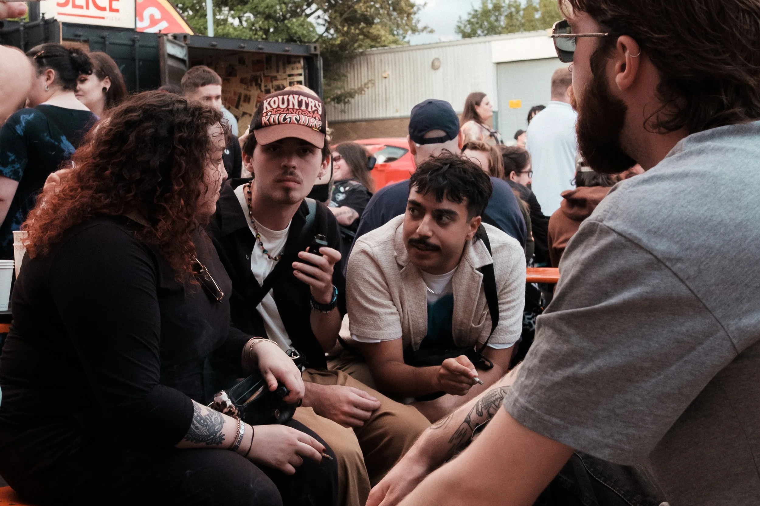 Group of young adults sitting outdoors at a social gathering or festival, engaged in conversation. There are food trucks and trees in the background.