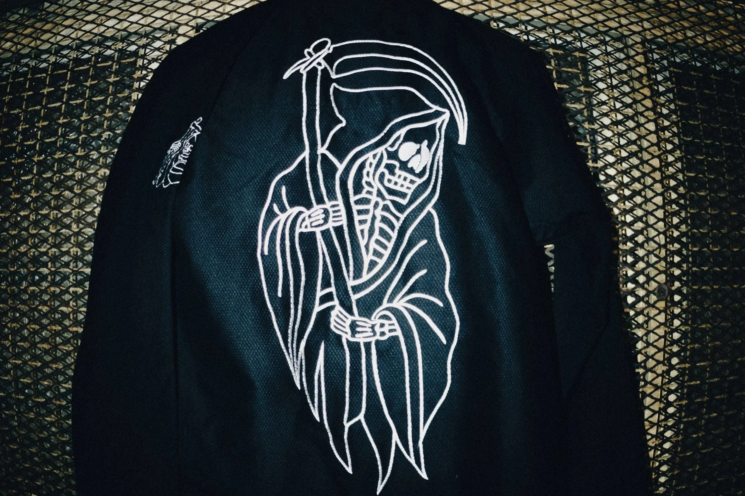 A black jacket with a white embroidered skeleton figure dressed as a Grim Reaper, holding a scythe, hanging on a metal mesh rack.