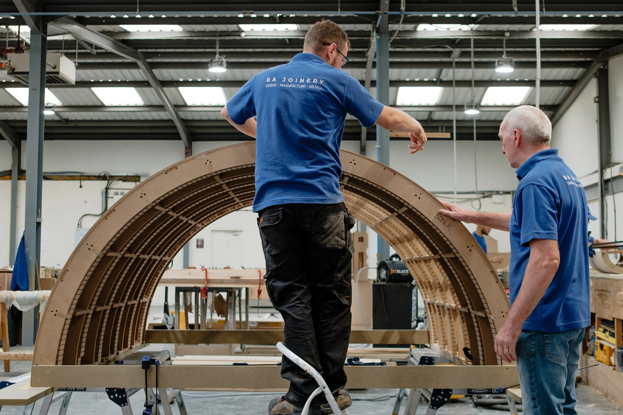 Two workers assembling a large wooden arc in a workshop, with various tools and stacks of wood around them.