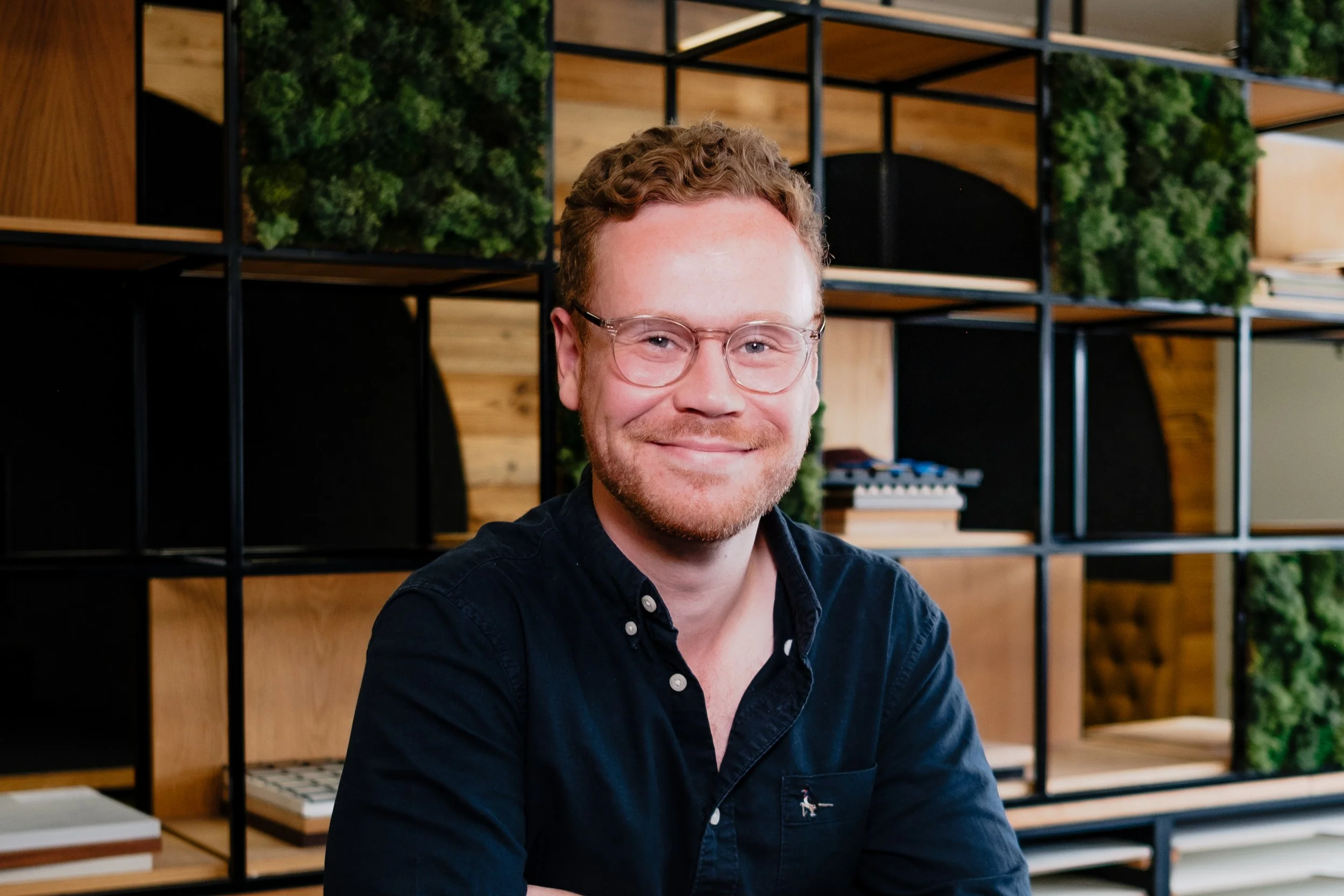 A man with glasses and a beard sitting at a table with a white mug, smiling at the camera in a modern office or coworking space, with a decorative wooden and green plant shelving unit behind him.