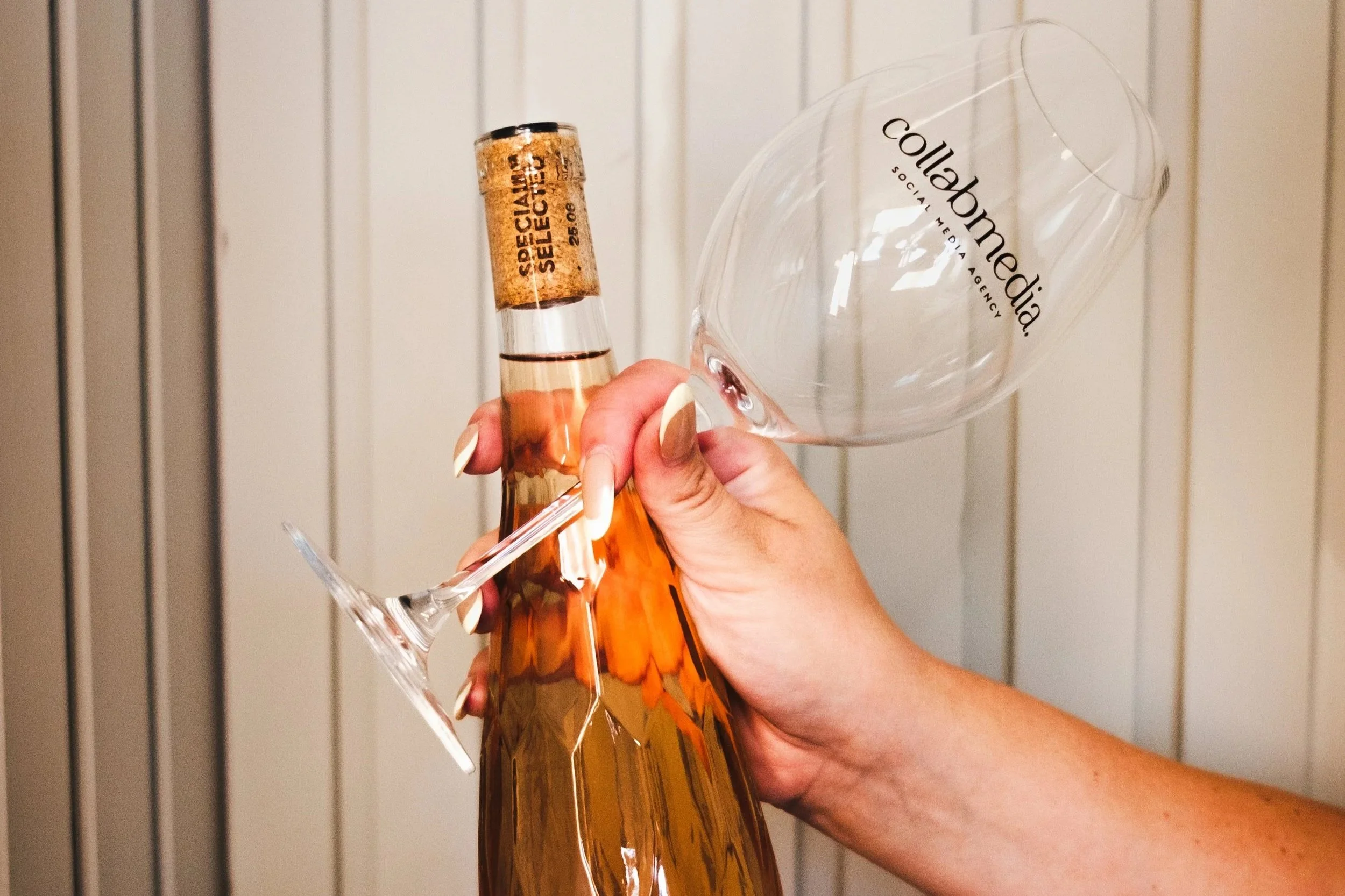 A hand holding a bottle of rosé wine with a wine glass balanced upside down on the neck of the bottle, against a vertical paneled background.