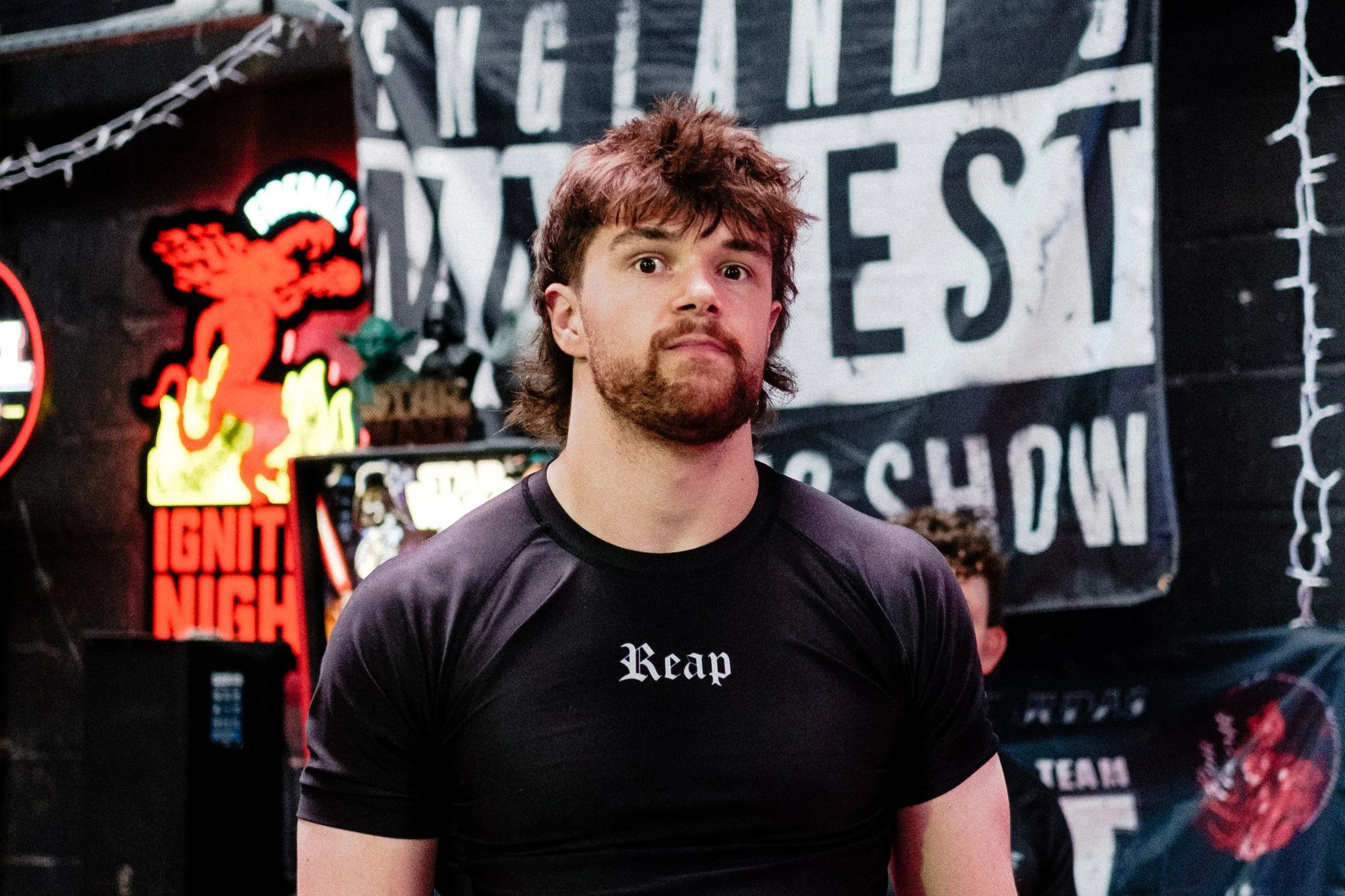 A man with brown hair and a beard wearing a black T-shirt with the word 'Reap' standing inside a store with dark walls, various neon signs, and posters including a Star Wars themed one in the background.