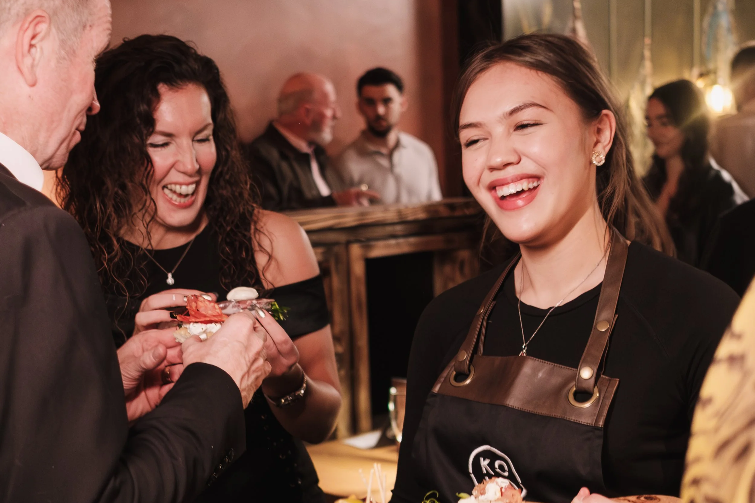 A young woman wearing a black apron serving food and drinks on a wooden tray at a social gathering or party, smiling and engaging with guests.