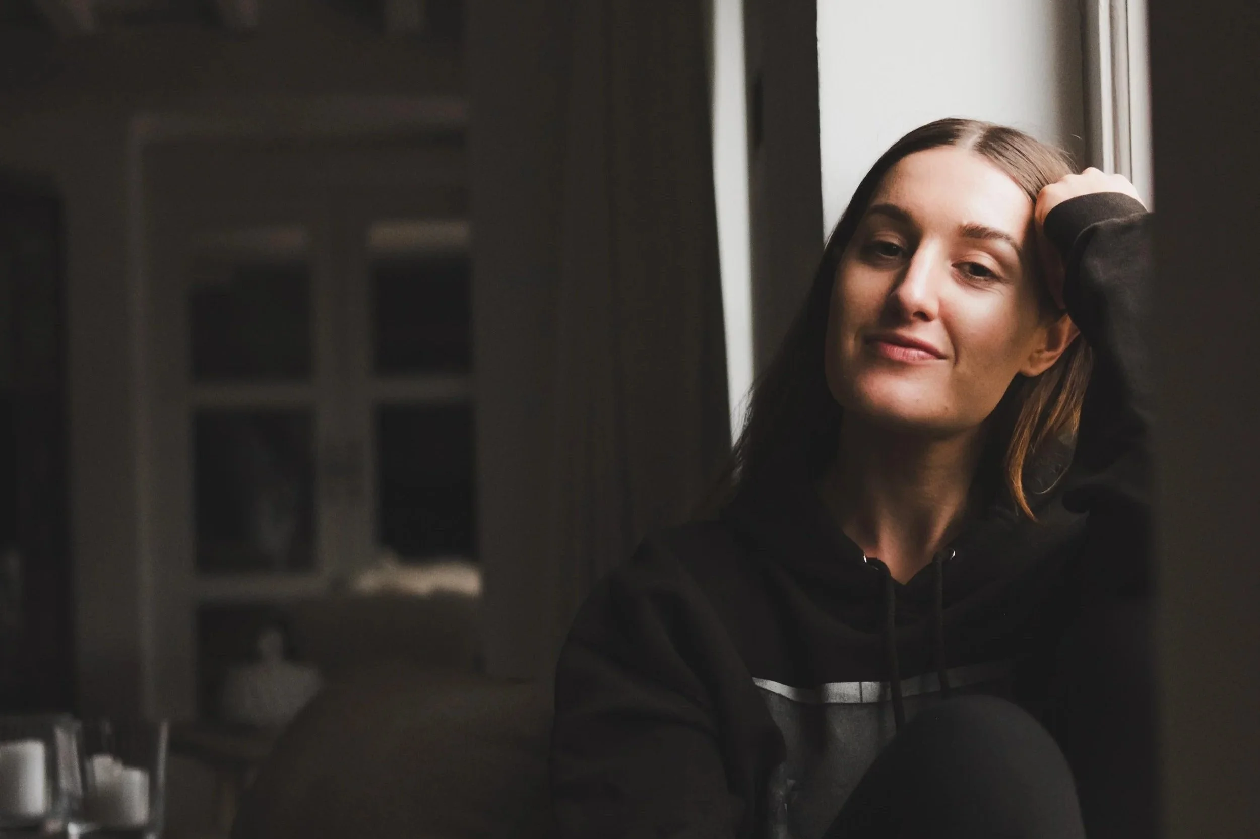 A young woman with long brown hair, wearing a black hoodie, sitting by a window with her hand on her head, smiling slightly, in a dimly lit indoor setting.