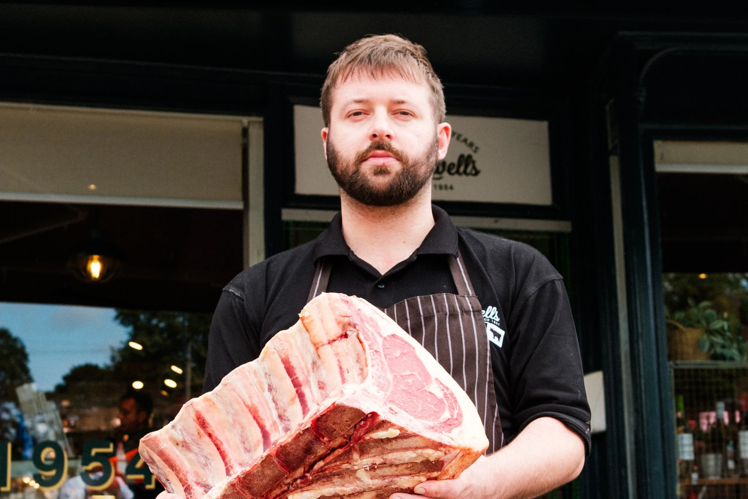 Man wearing an apron holding a large slab of raw bacon outside a storefront.