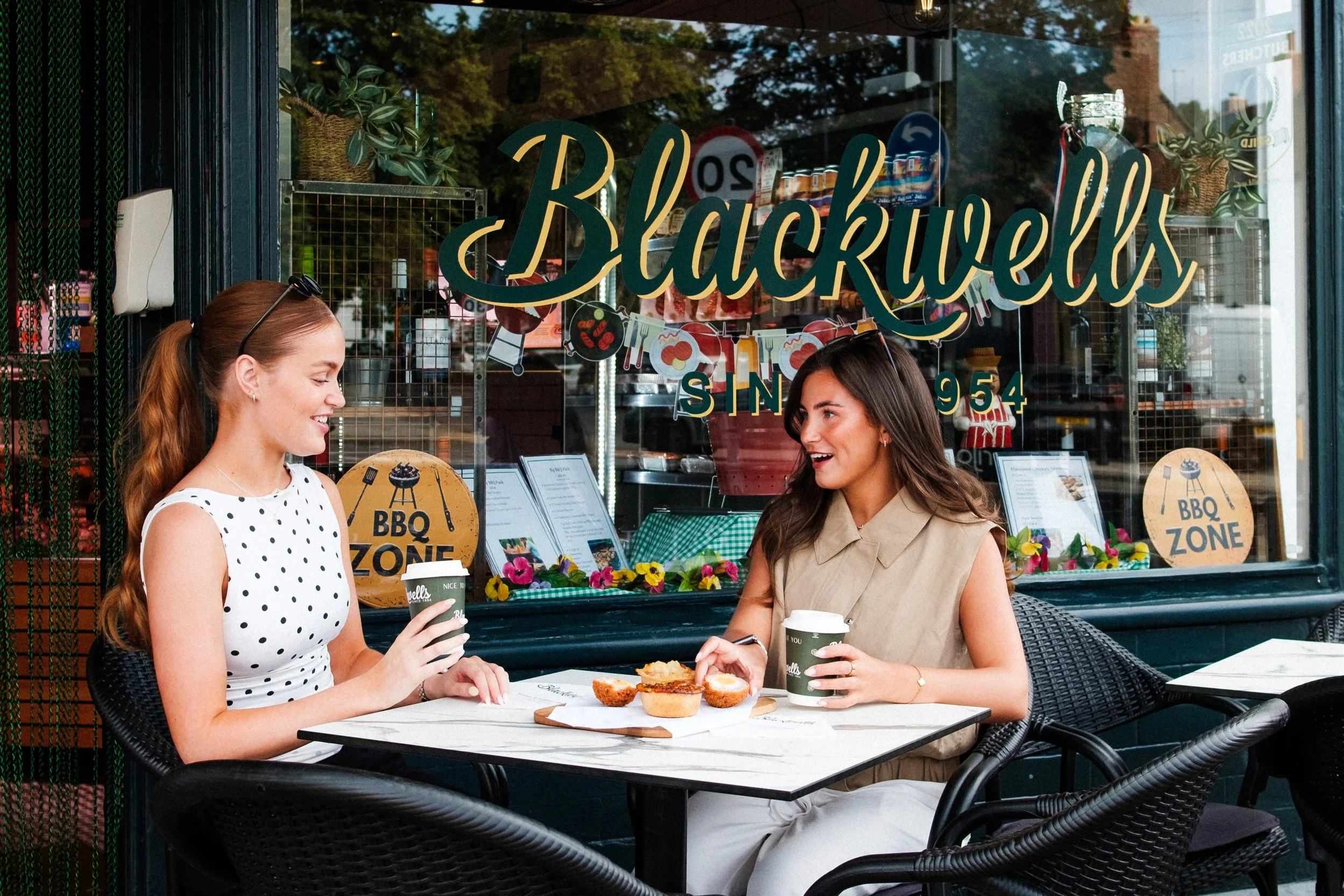 Two women sitting at an outdoor cafe table in front of a store window named Blackwells, enjoying coffee and pastries, with signs advertising a BBQ zone inside.