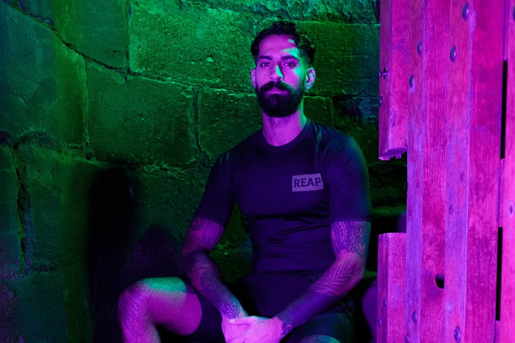 A bearded man with tattoos sitting in a small, colorful room with green and purple lighting. He is wearing a black shirt with "REAP" on it, shorts, striped socks, and sneakers.