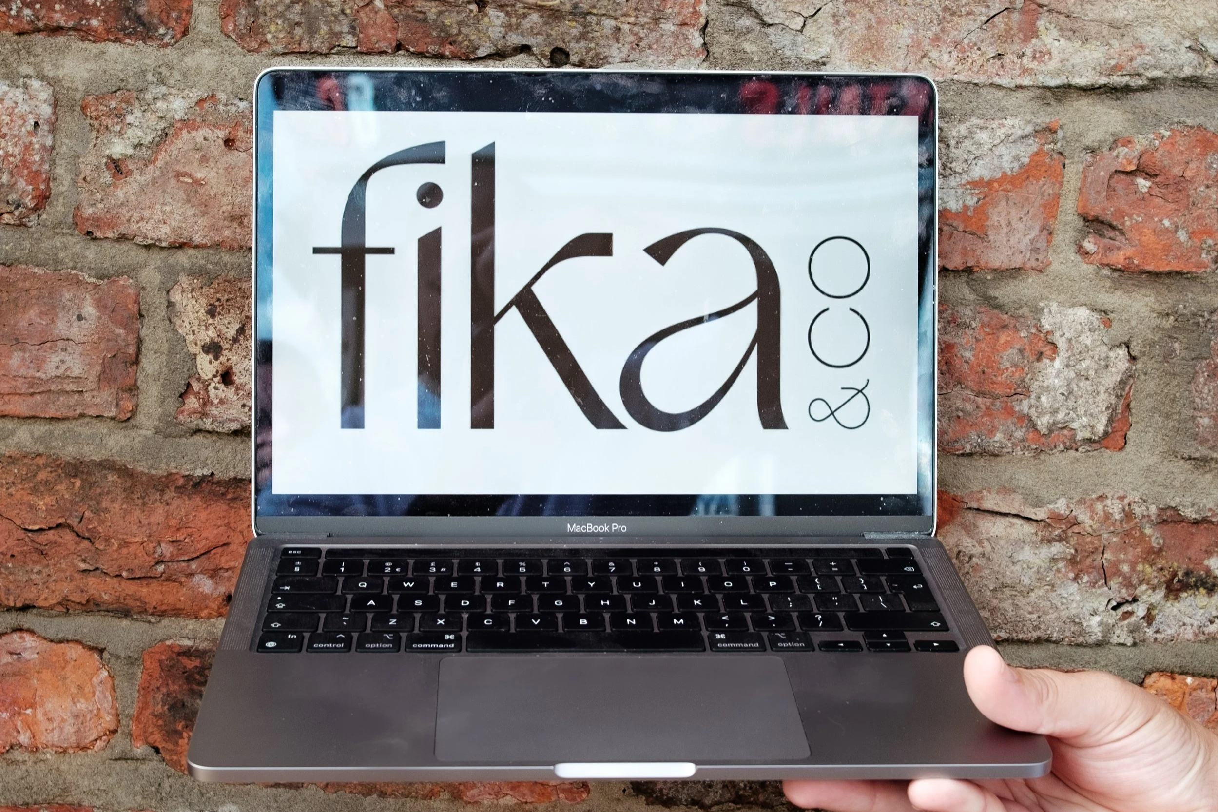 A person holding a MacBook Pro laptop with a screen displaying the word 'fika' against a brick wall background.