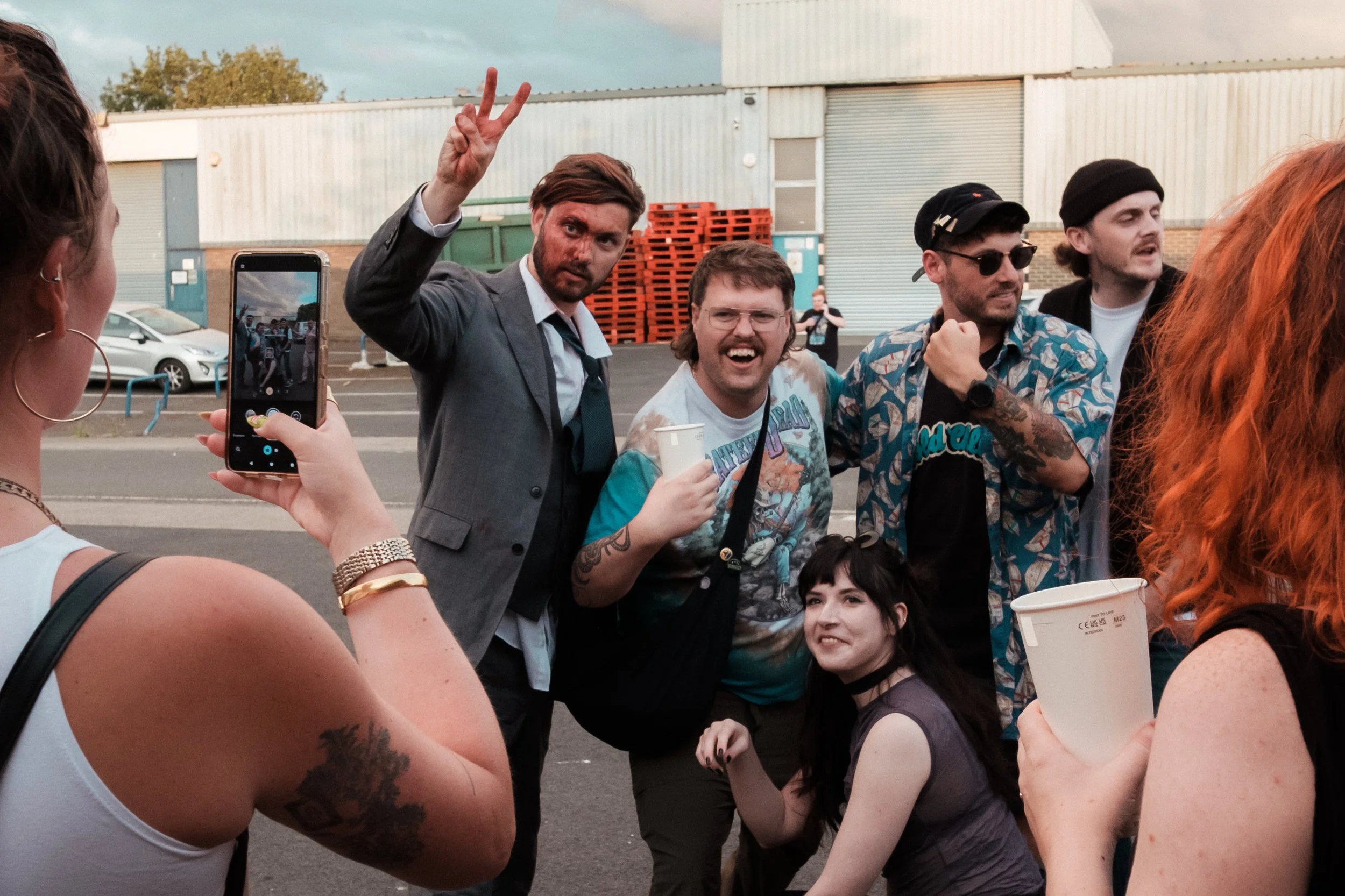 Group of young friends taking a photo together outdoors, some making peace signs and smiling, in a parking lot with a warehouse behind them.