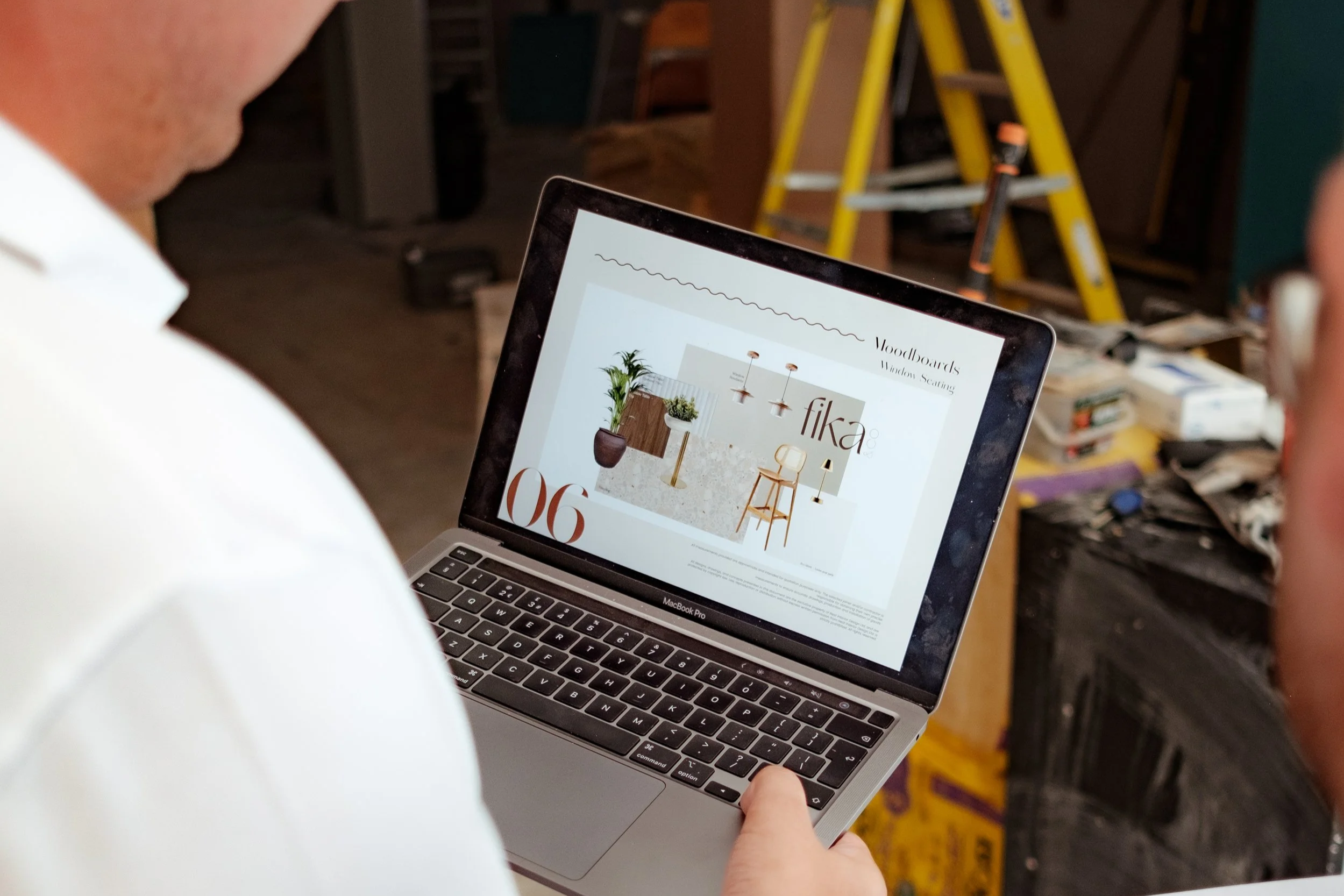 Person holding a laptop showing a design catalog with the title 'Fika' in a workspace with a ladder and tools in the background.