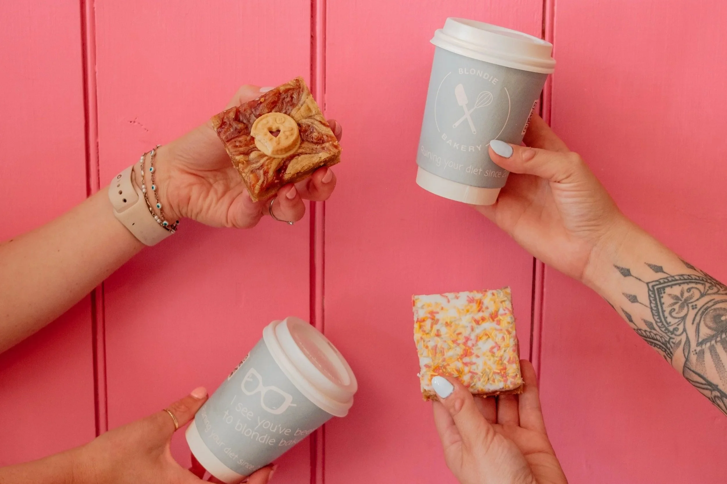 Three hands holding different snacks and coffee cups against a pink wooden background.