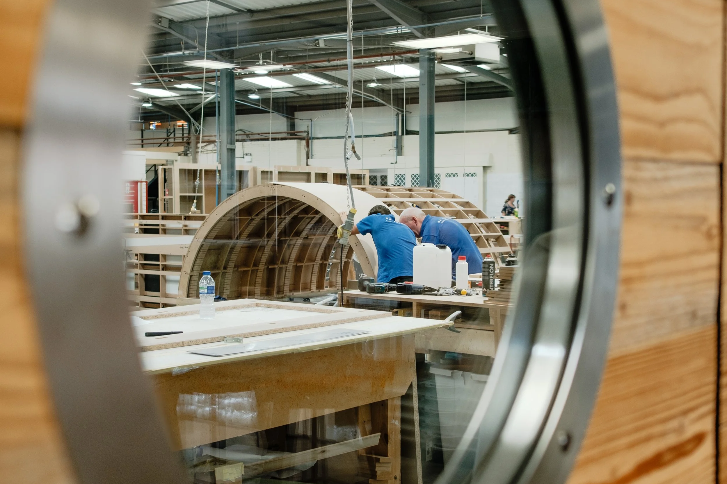 View through a circular window showing two people working on a wooden boat inside a workshop.