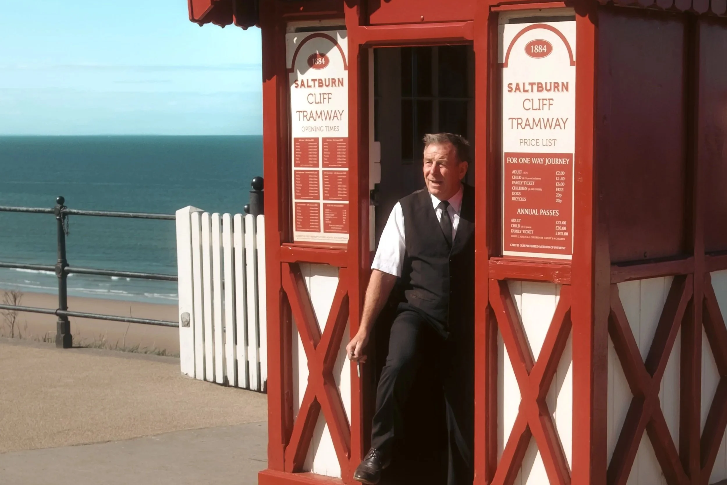 A man dressed in formal attire standing inside a red old-fashioned tram kiosk by the seaside with the ocean in the background