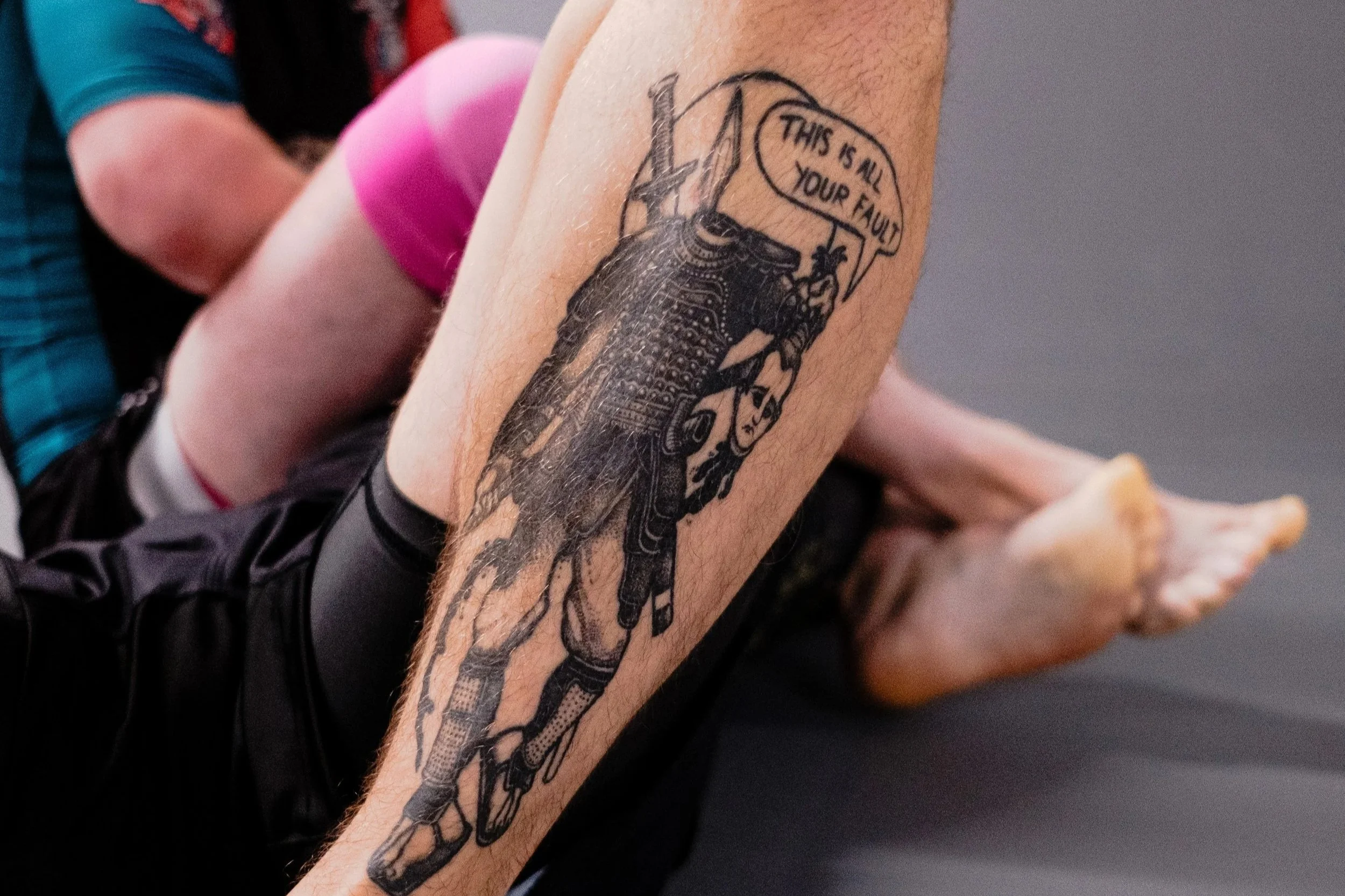 Close-up of a tattoo on a person's leg featuring a warrior figure with a speech bubble that says, 'This is all your fault.'