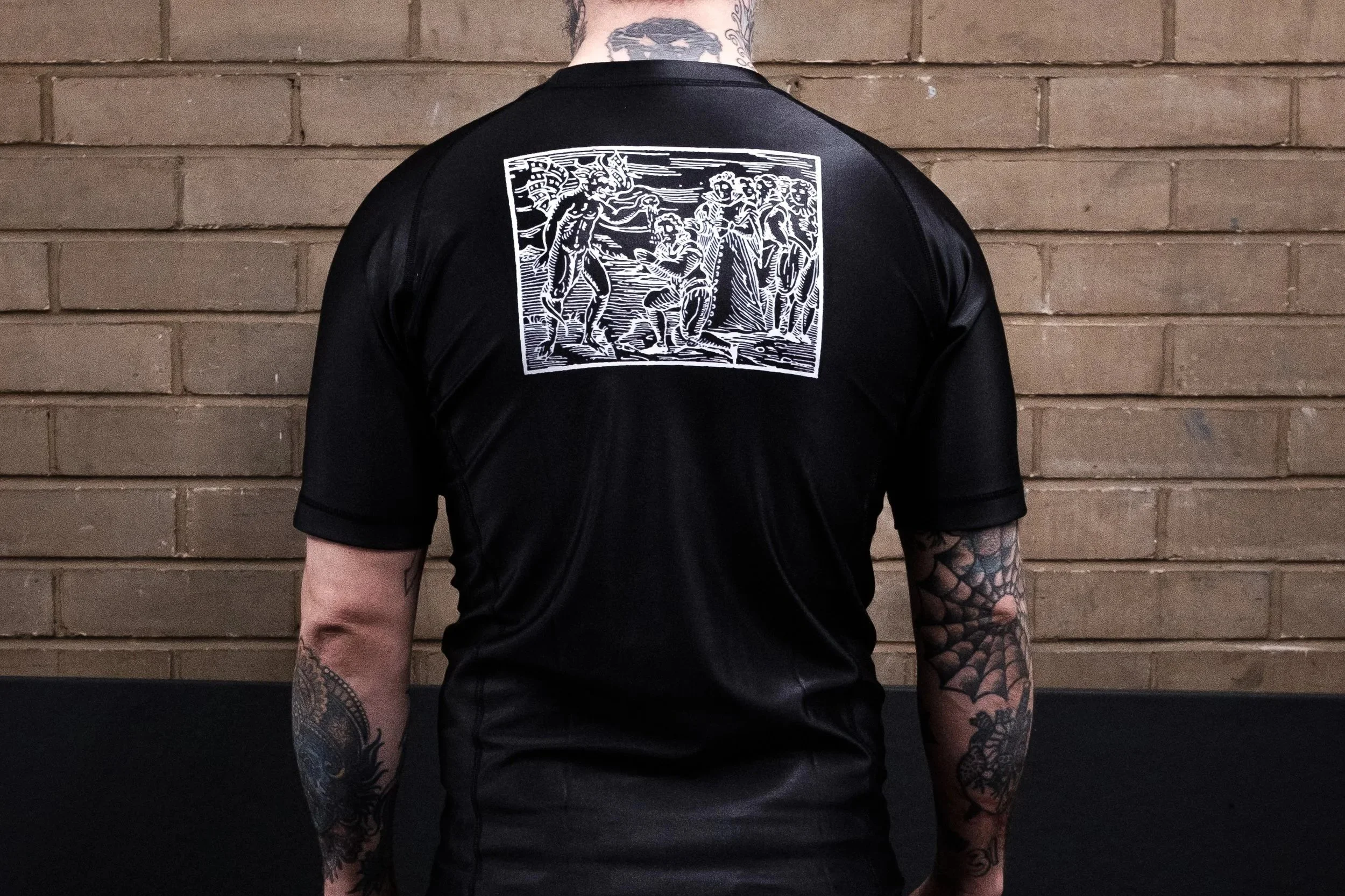 Back view of a person wearing a black sports jersey and black shorts, standing against a brick wall, with tattoos on their arms and a tattoo on their leg.