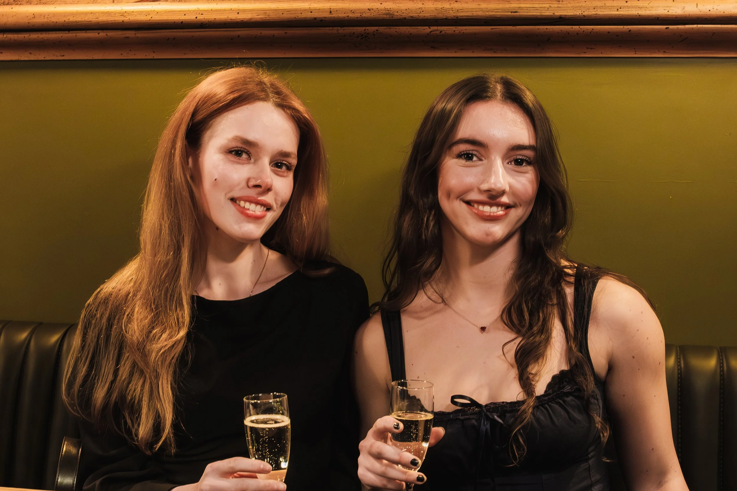 Two young women sitting at a bar or restaurant, smiling and holding glasses of champagne.
