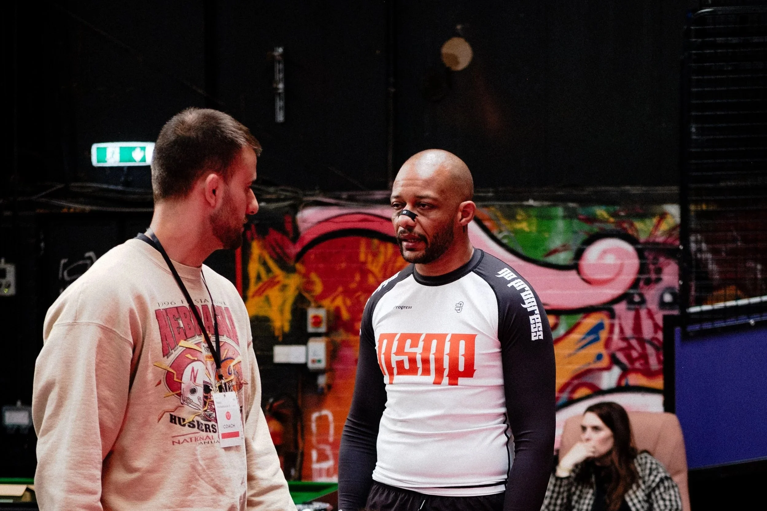 Two men with athletic builds and tattoos, one sitting cross-legged on a pink mat and the other standing, are having a conversation in a gym. The background features a colorful graffiti wall, a woman sitting in a chair watching, and gym equipment.
