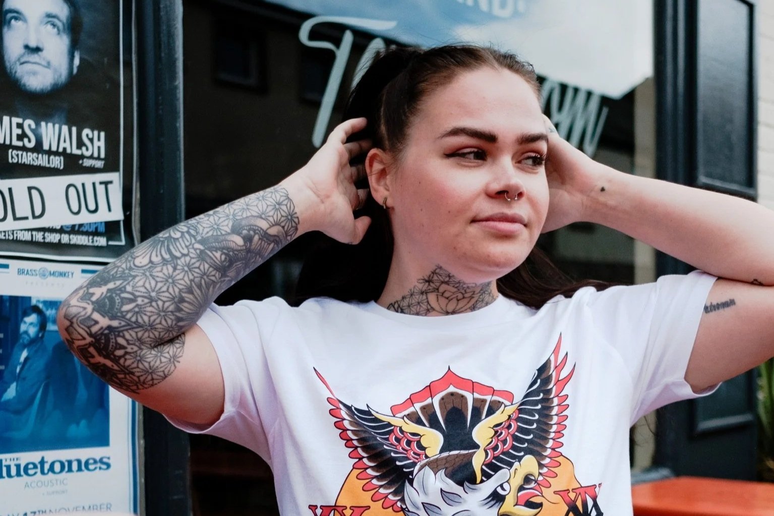 A young woman with tattoos on her arms and neck, wearing a white graphic T-shirt, sitting at an outdoor table. She is adjusting her hair with her hands. There are orange tables and glasses in the foreground, with posters and a window behind her.