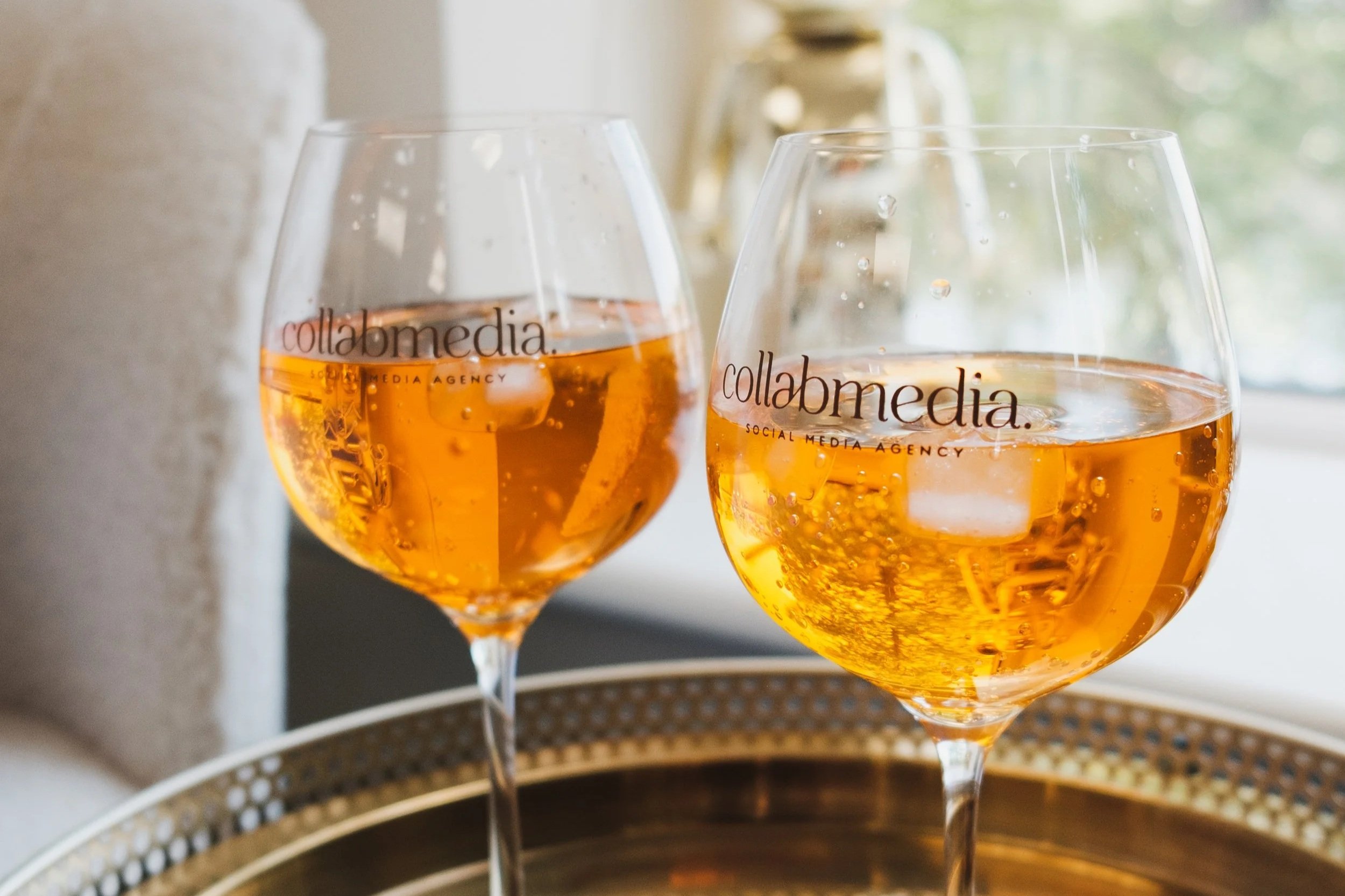 Two glasses of rosé wine on a gold tray, with a white phone, a gold pen with a pineapple top, and a gold ring, near a window.