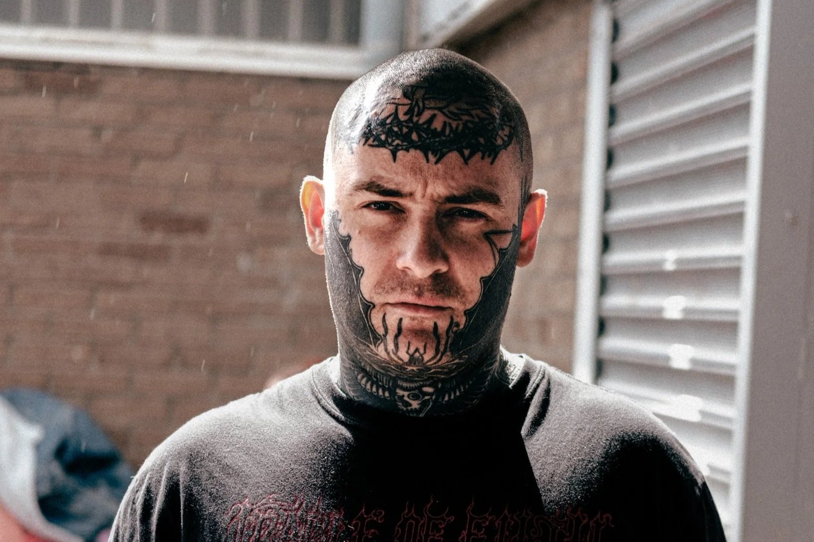 A man with tattoos on his face, head, and arms holding two cups of beer, standing outside near a brick wall and a metal siding door.
