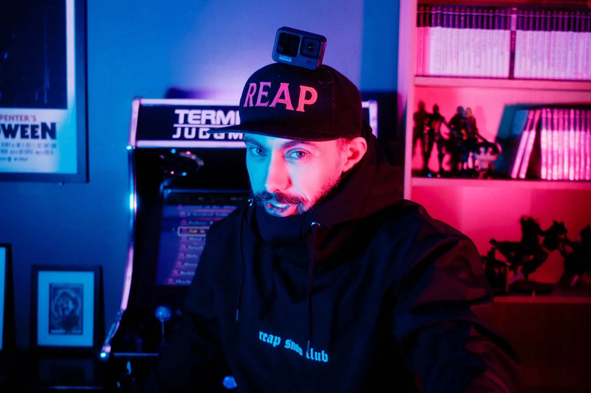 A man wearing a black hoodie and cap with "REAP" on it, with a GoPro camera on his cap, sitting in a room with neon pink and blue lighting, surrounded by shelves with action figures and books, and an arcade machine in the background.