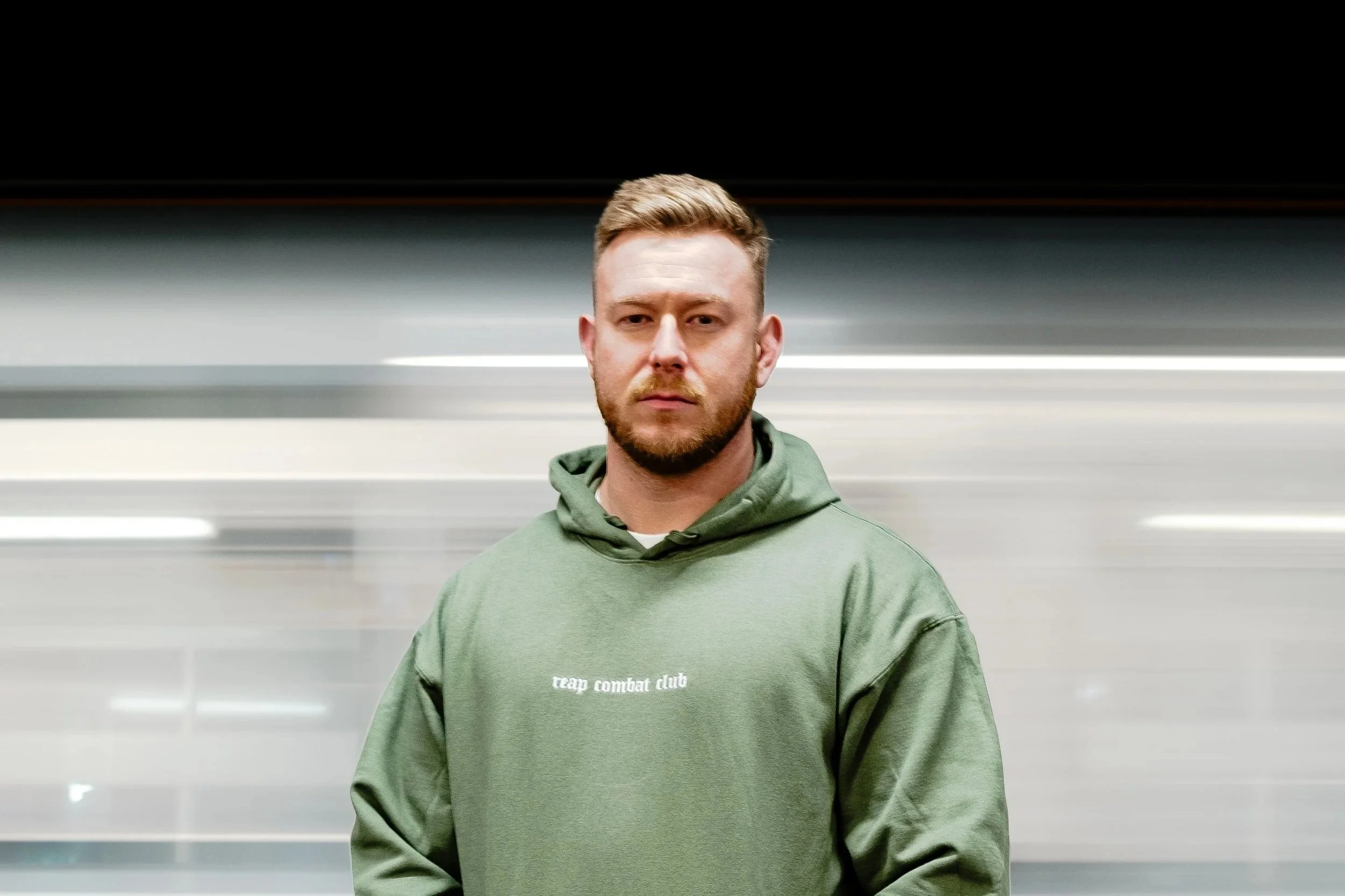 A man in a green hoodie stands in front of a blurred background, looking directly at the camera with a serious expression.