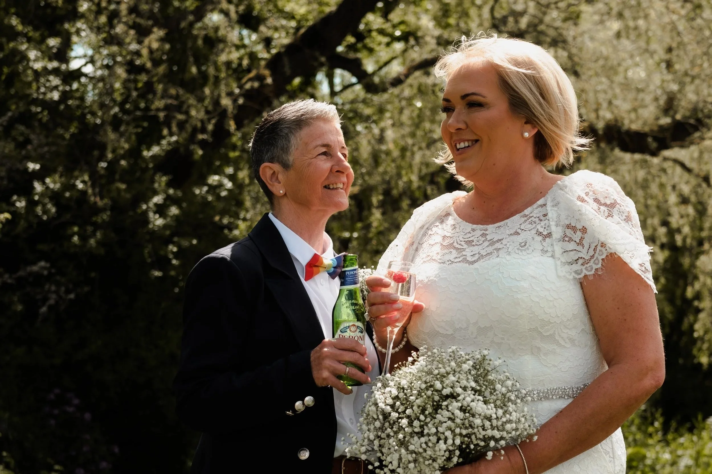 Two women smiling and chatting outdoors during a wedding celebration. One woman is wearing a black blazer, beige pants, and a colorful bow tie, holding a drink. The other woman is dressed in a white lace wedding gown, holding a bouquet of white flowe
