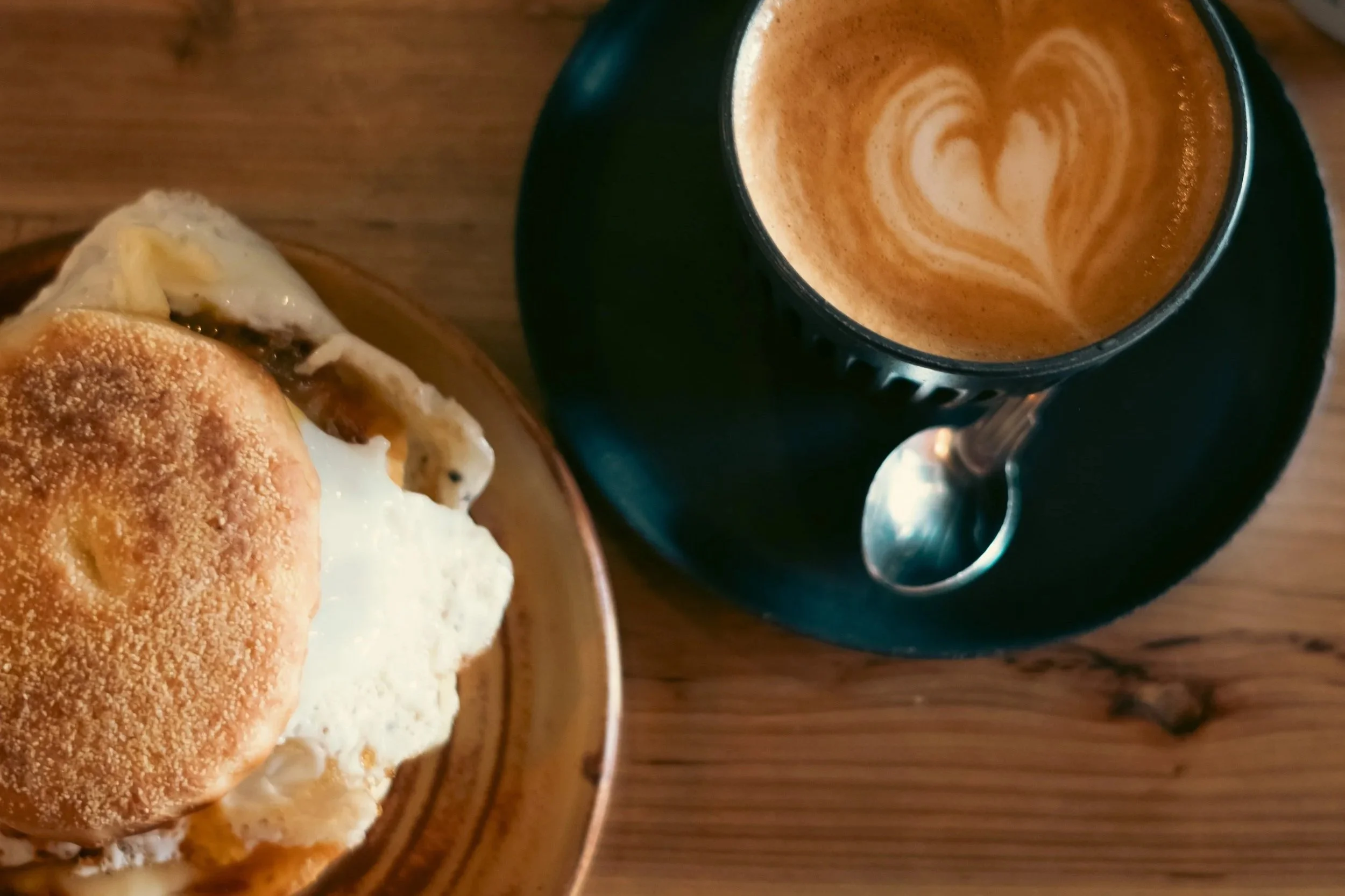 A cup of coffee with latte art in a black mug and a small plate with a biscuit sandwich on a wooden table.