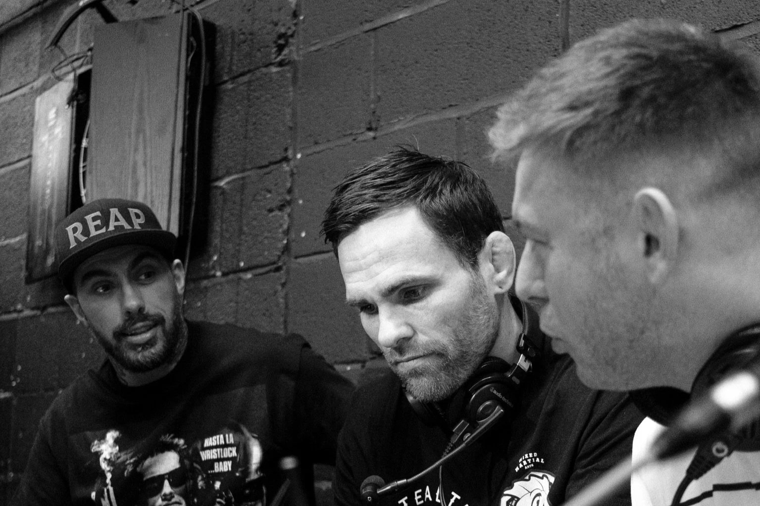 Three men sitting at a table with audio equipment in front of them, engaged in a discussion against a brick wall background.