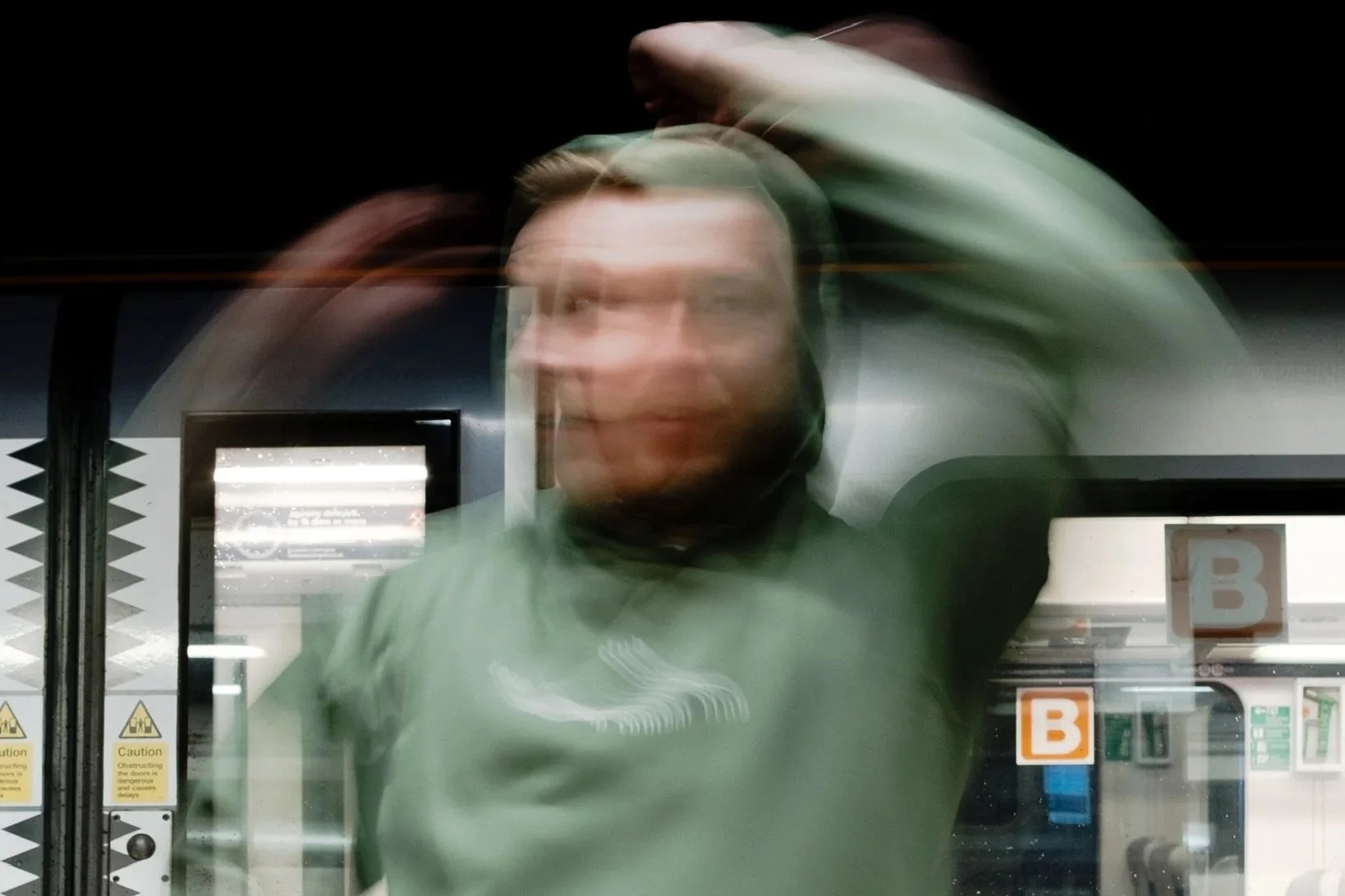 Person in a green hoodie adjusting their hood in front of a subway train door with the letter B signs beside it