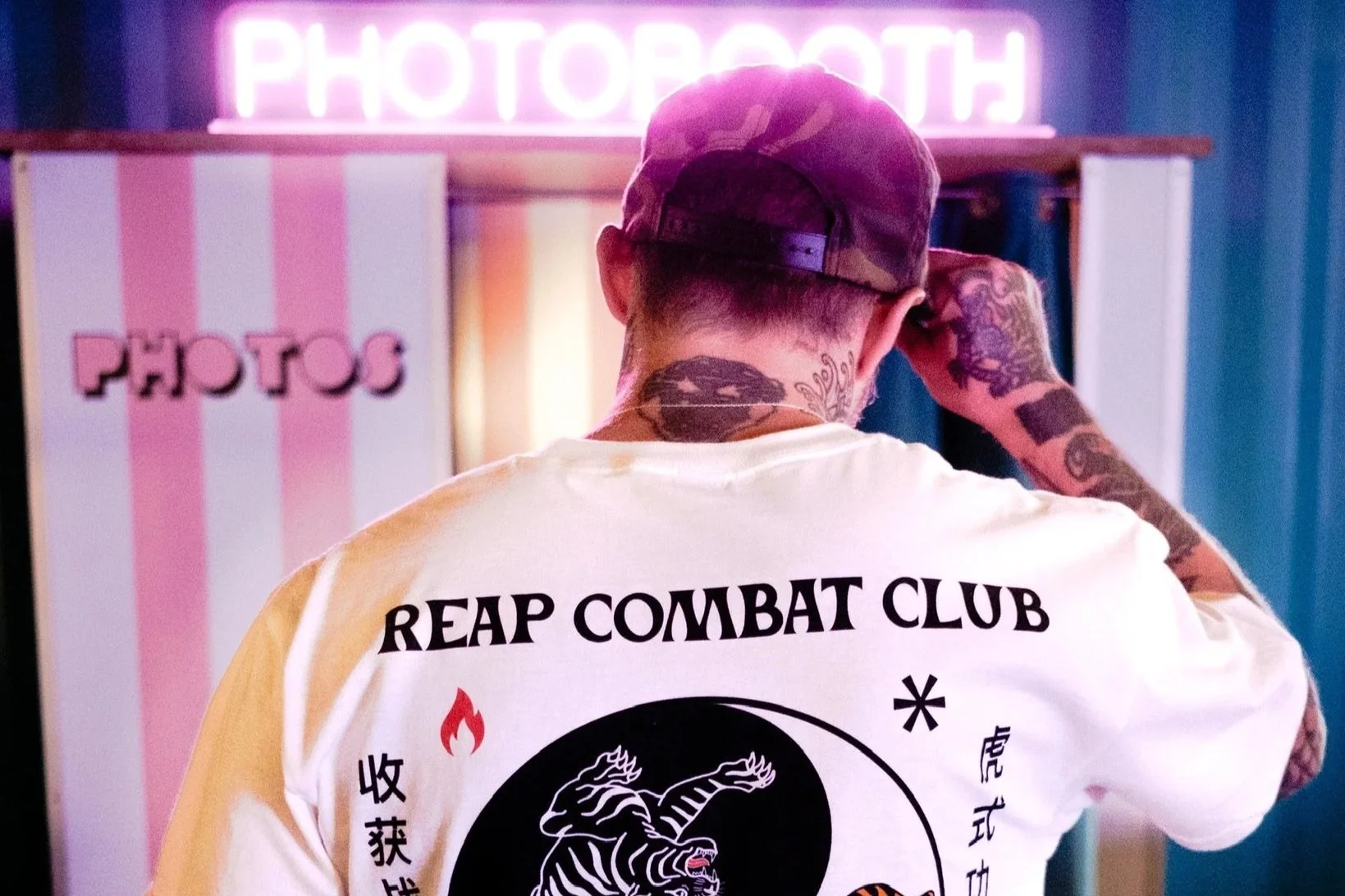 Back of man wearing a white t-shirt with a tiger and dragon illustration, and the text 'REAP COMBAT CLUB' and 'Tiger Style Jiu Jitsu - No Mercy'. The man is in front of a photo booth with a neon sign that says 'PHOTOBOT' and a pink and white striped 
