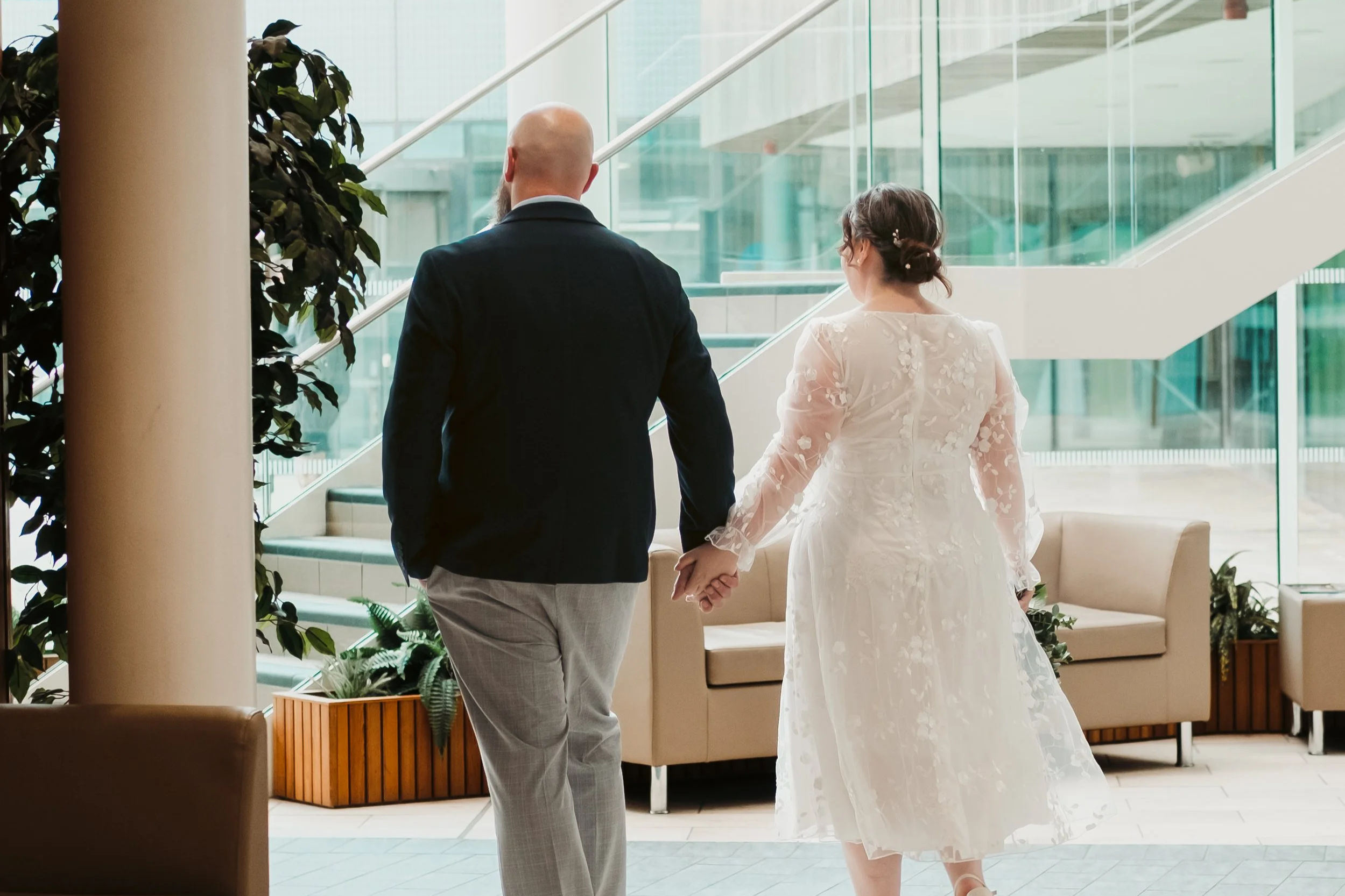 A couple holding hands walking inside a modern building with large glass windows, green plants, and comfortable seating.