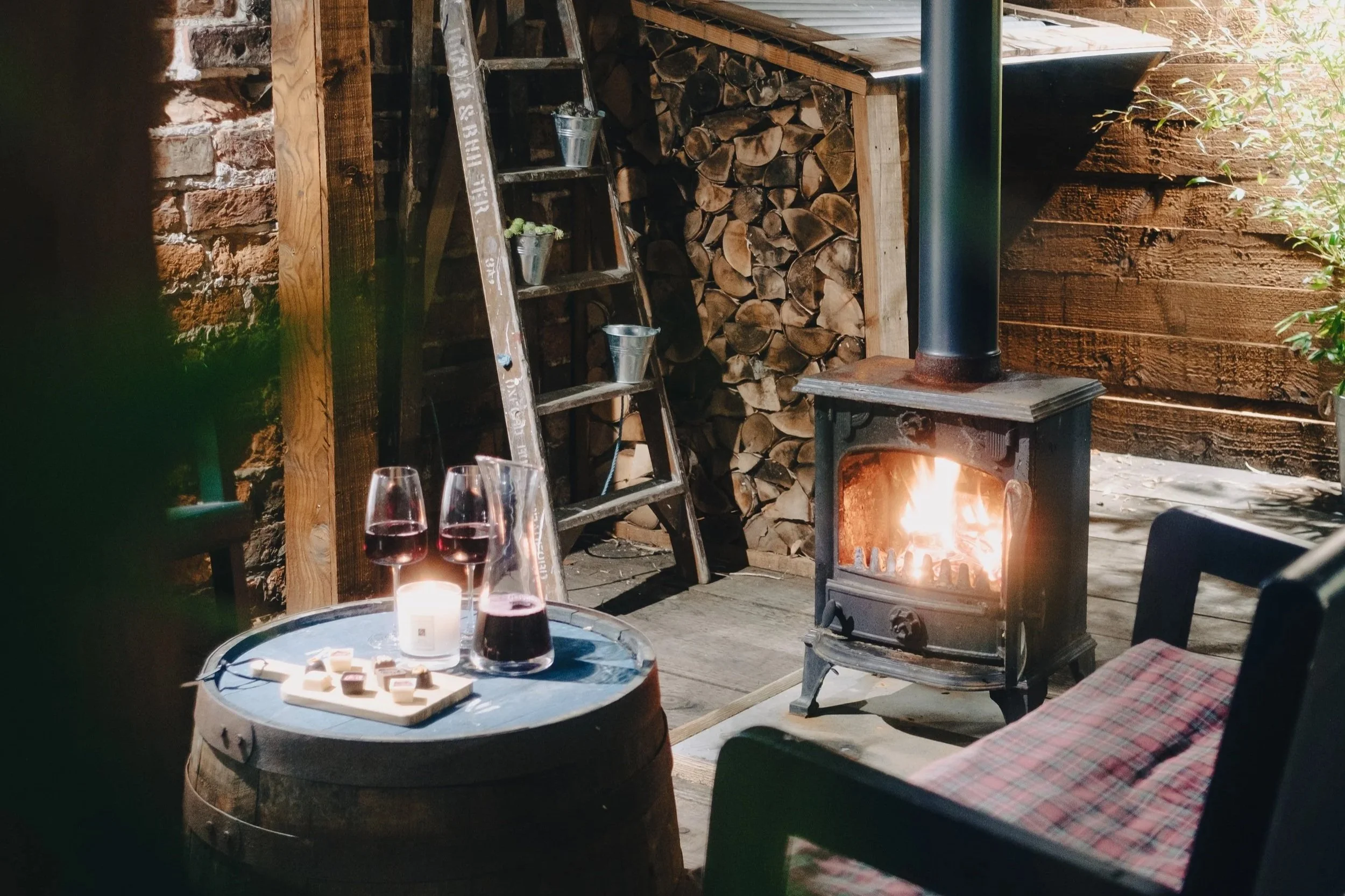 Cozy outdoor patio with a wood-burning stove, a small table with three glasses of red wine, a candle, and a tray of snacks. There is a ladder, split firewood, and greenery in the background.