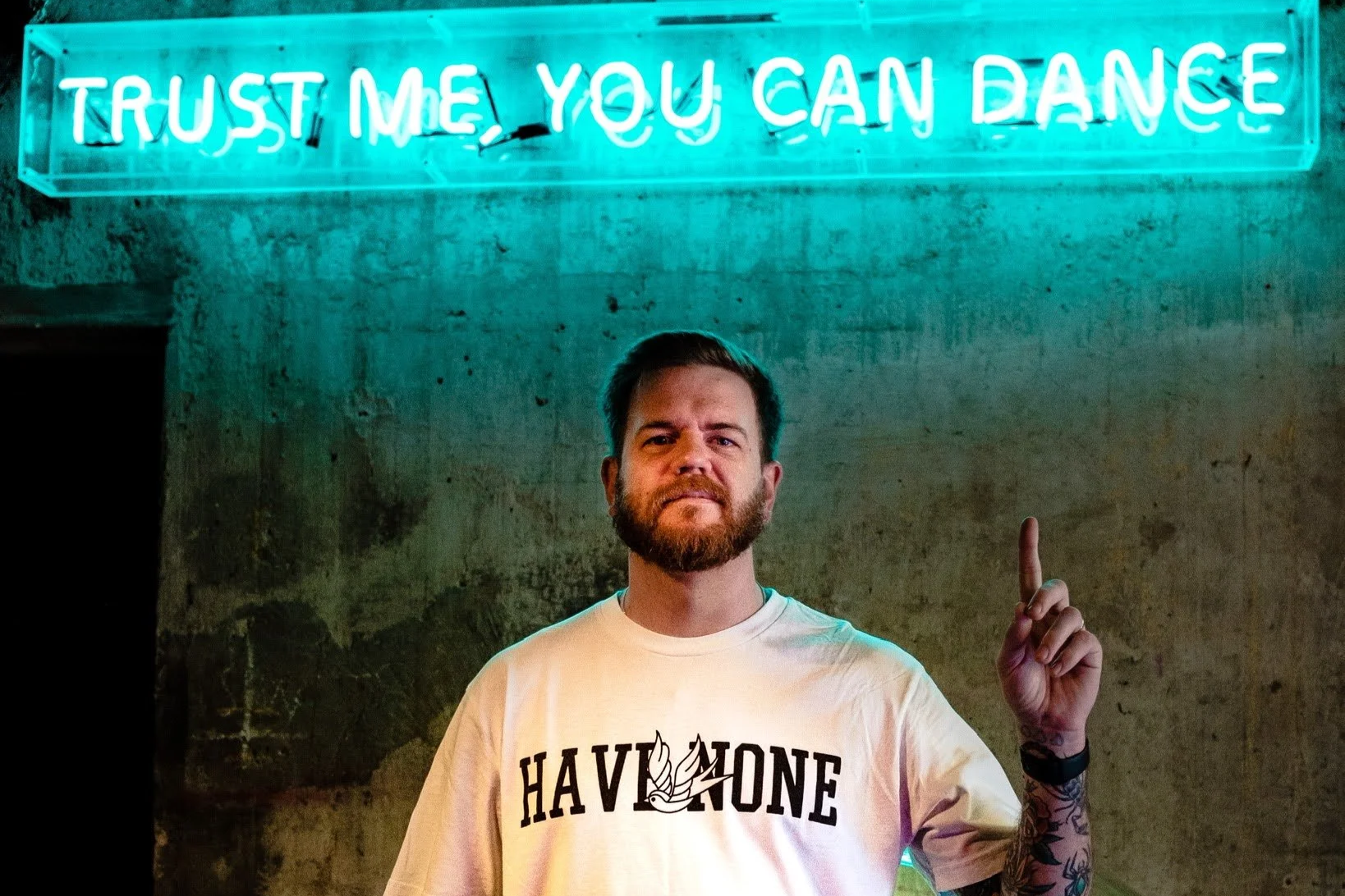 A man with a beard and tattoos standing in front of a neon sign that reads "Trust Me, You Can Dance." He is wearing a white T-shirt with the words "HAVE NONE" printed on it and is raising his right index finger.