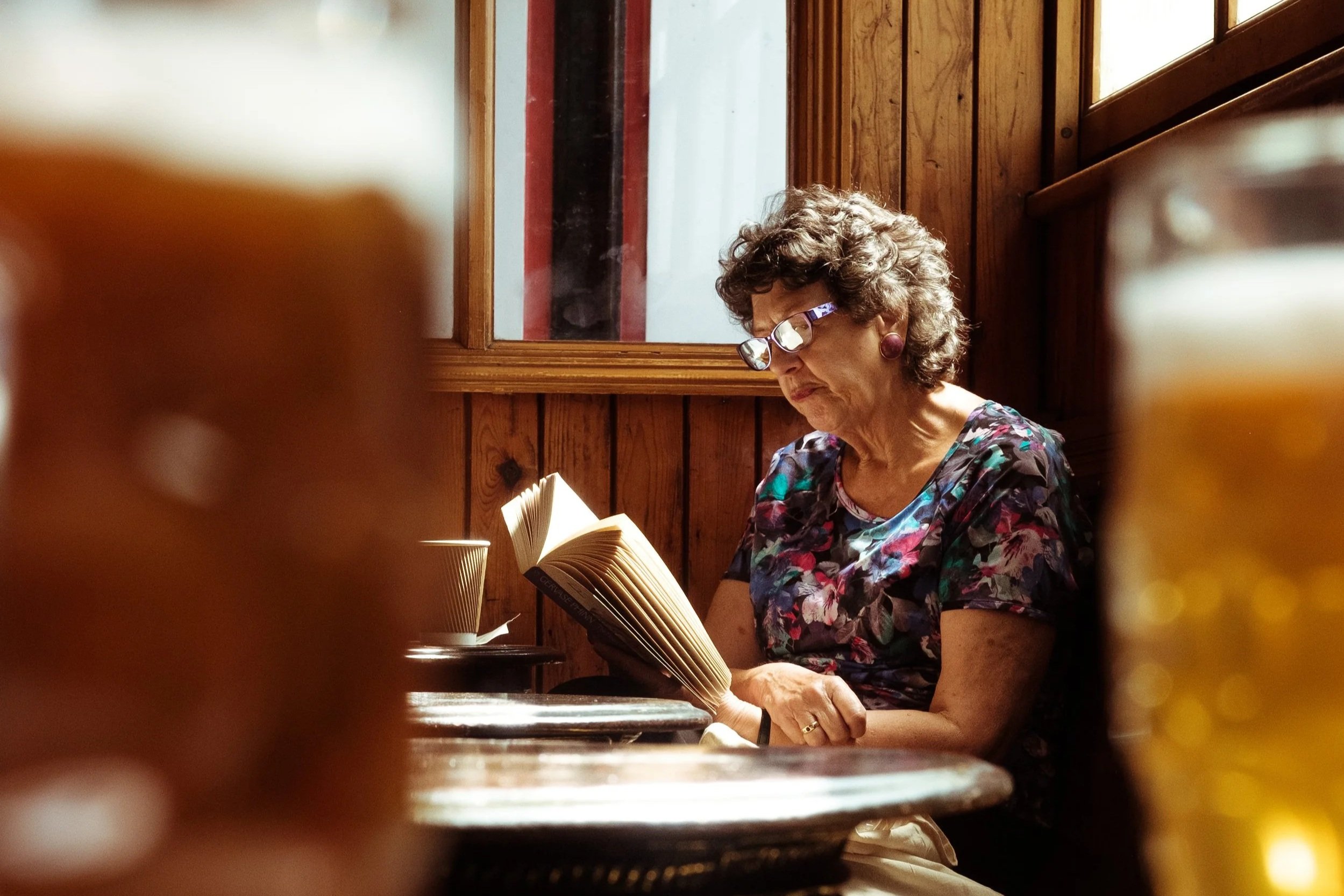 An elderly woman with curly hair and glasses reading a book in a rustic café with wooden walls and windows.