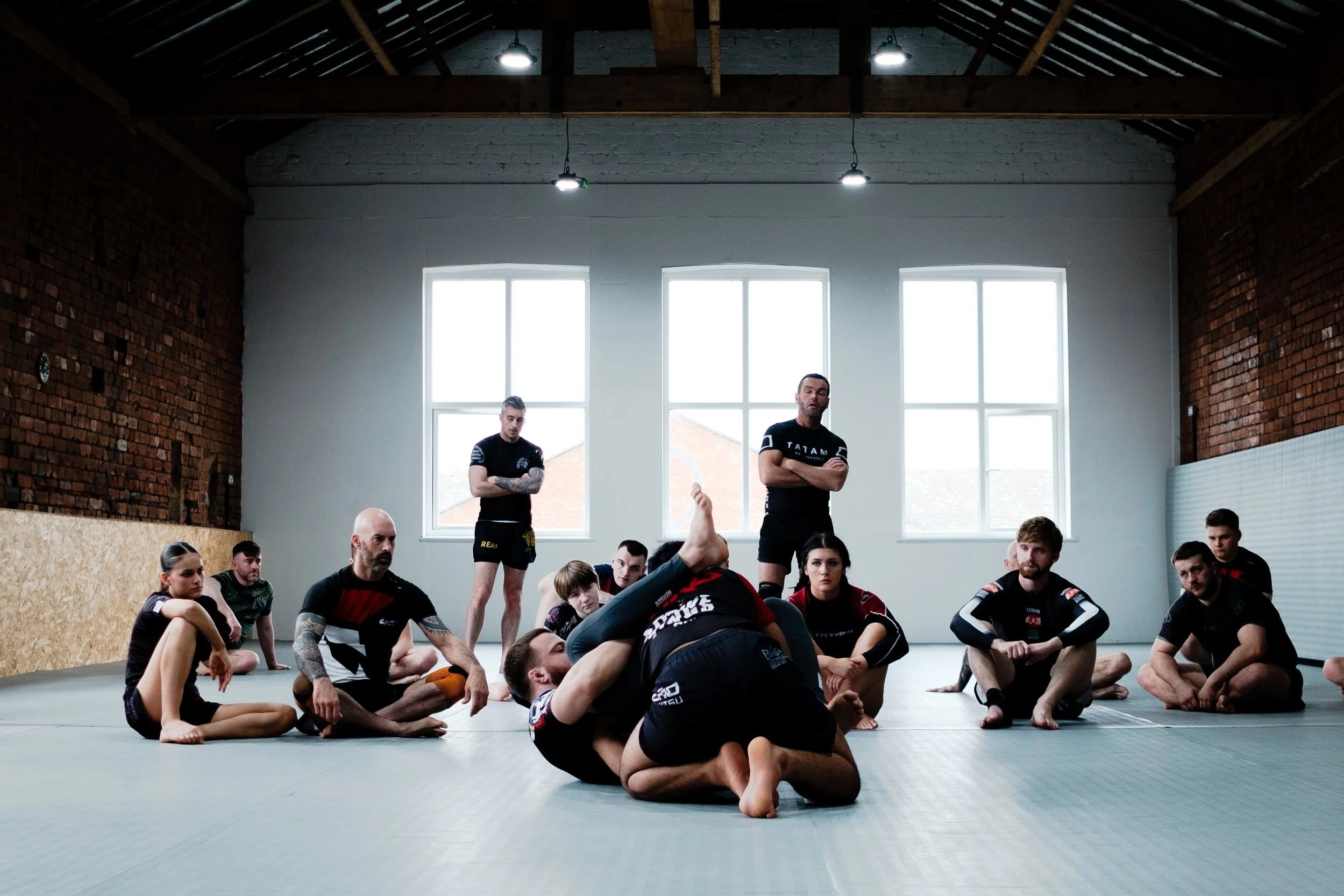 Mixed martial arts class training in a spacious gym with wooden beams and large windows, with students practicing grappling techniques on mats.