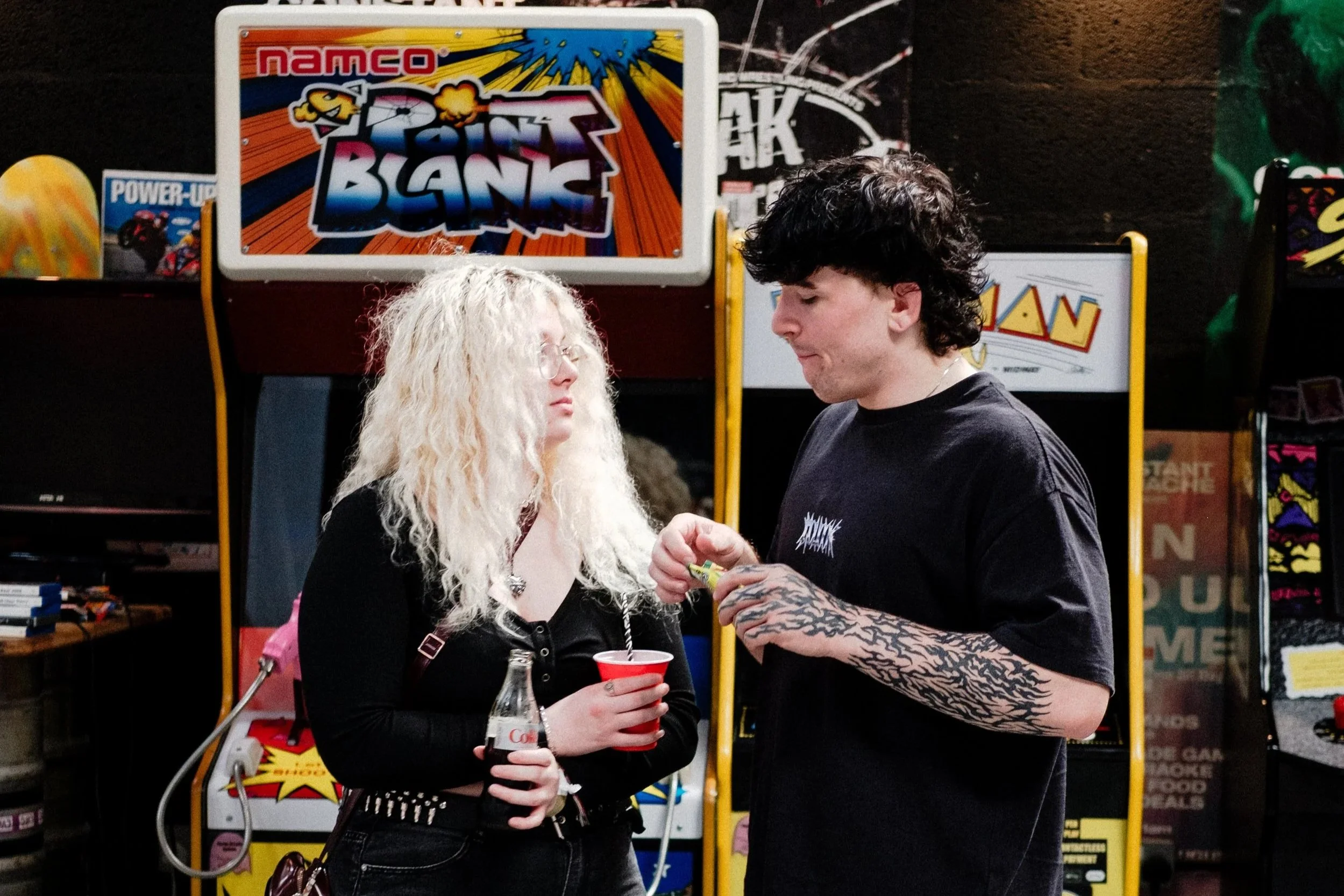 A young woman and a young man are standing face to face in front of arcade machines, engaging in conversation. The woman has long, curly blond hair, glasses, and is holding a red cup and a bottle of Coca-Cola. The man has black, wavy hair, tattoos on