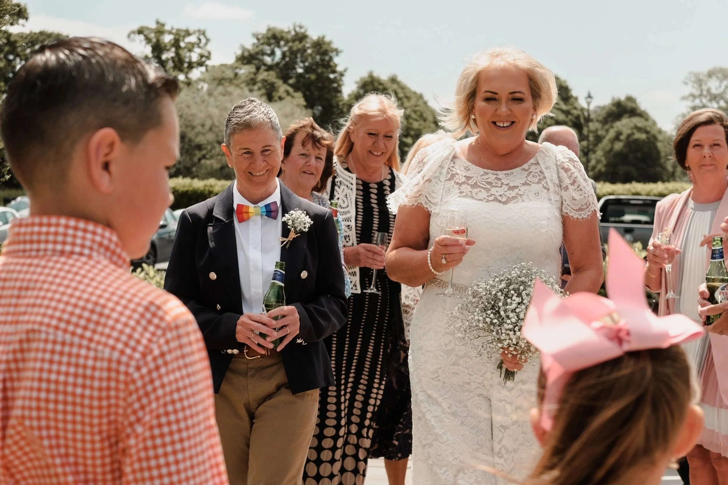 A group of people, including a woman in a white lace dress holding flowers, gathered outdoors during a sunny day, celebrating at a wedding or special event.
