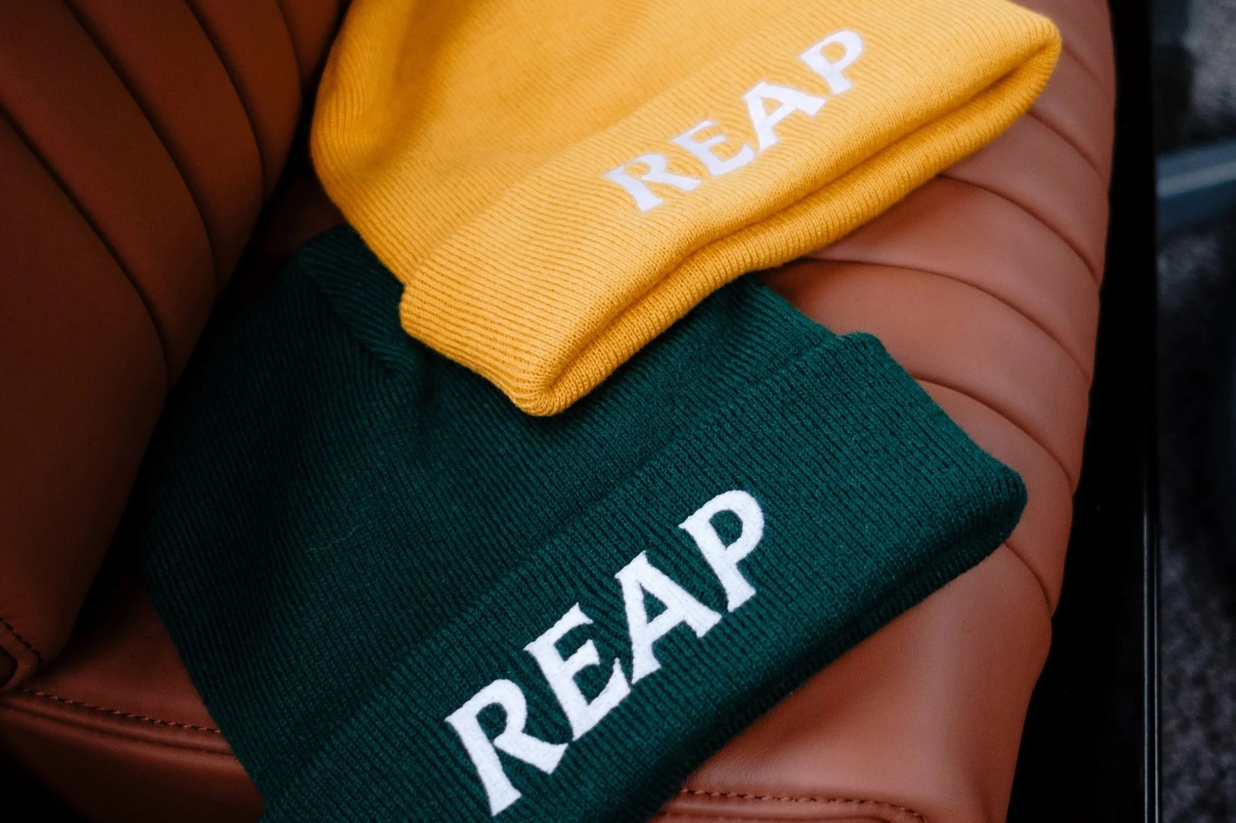 Yellow and green beanies with 'REAP' embroidered in white, placed on a brown leather seat.