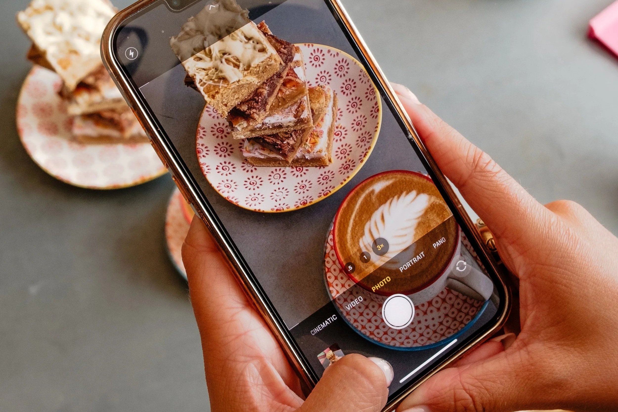 A person holding a smartphone taking a photo of plates of layered dessert bars and a latte on a gray table.