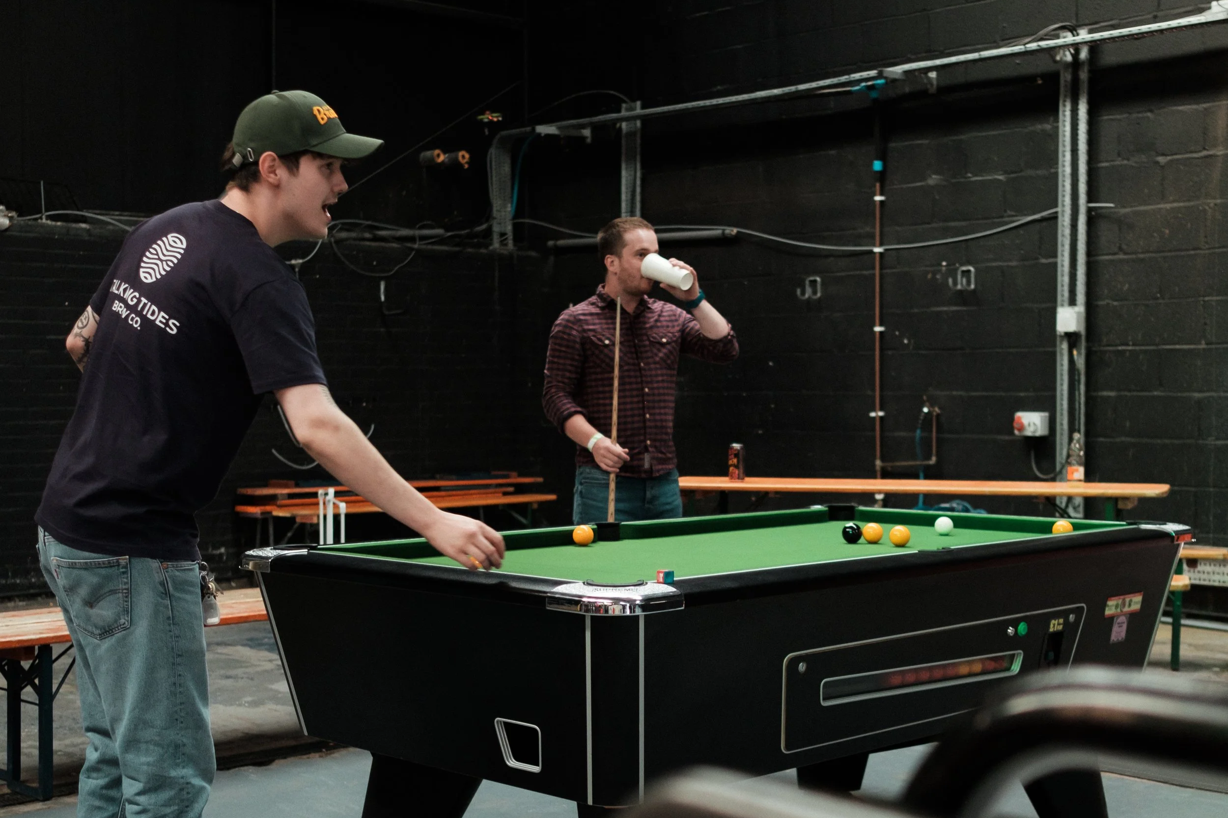 Two men playing pool in a game room with black walls and benches in the background.