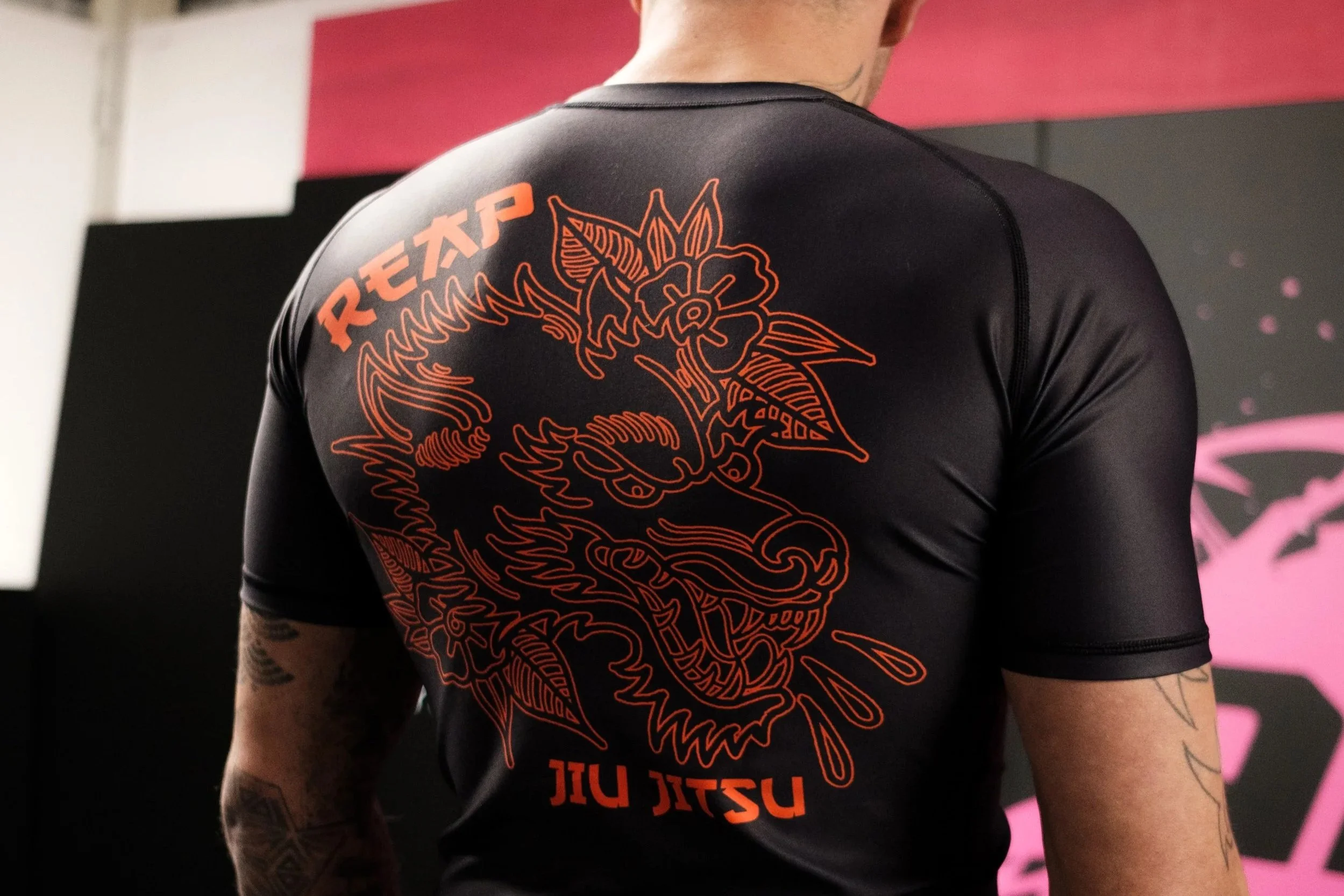 Back view of a person wearing a black rash guard with an orange dragon graphic and text, standing indoors in a martial arts gym.