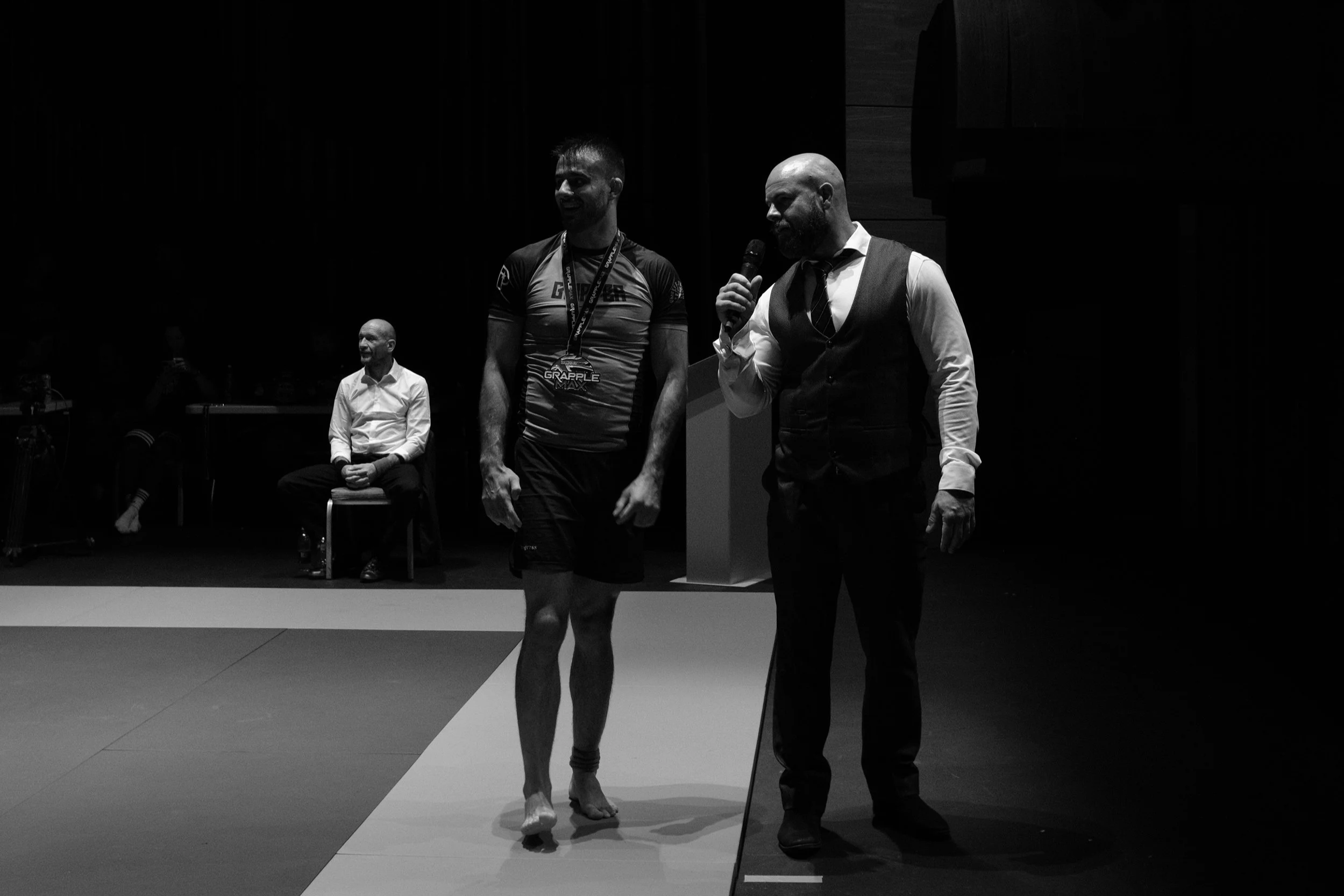Black and white photo of a man in athletic clothing walking across a mat while another man in a vest and tie speaks into a microphone. Two other men are seated behind them in a darkened room.