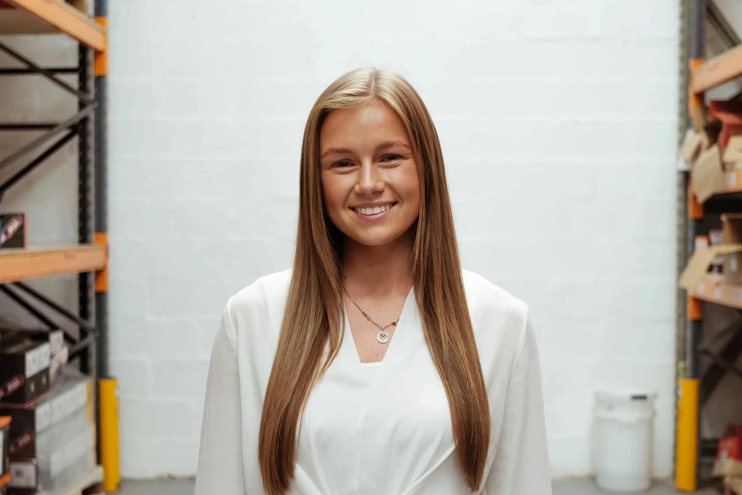 A young woman with long brown hair wearing a white blouse and black pants standing in a warehouse aisle with metal shelves on both sides filled with boxes and a white wall in the background.