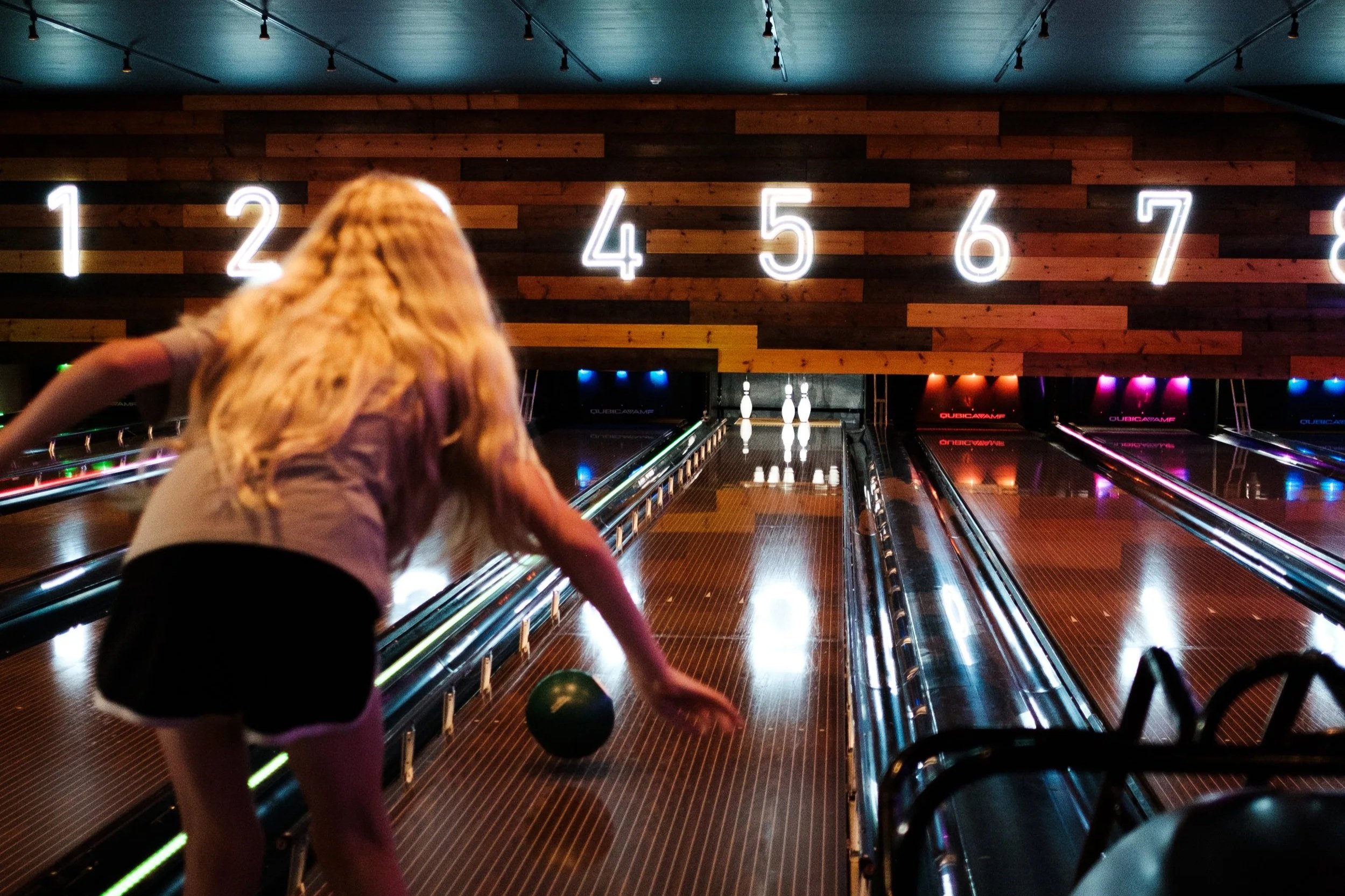 A girl bowling at an indoor alley, with glowing pins at the end of the lane and large illuminated numbers on a wooden wall behind. She is about to release a green bowling ball.