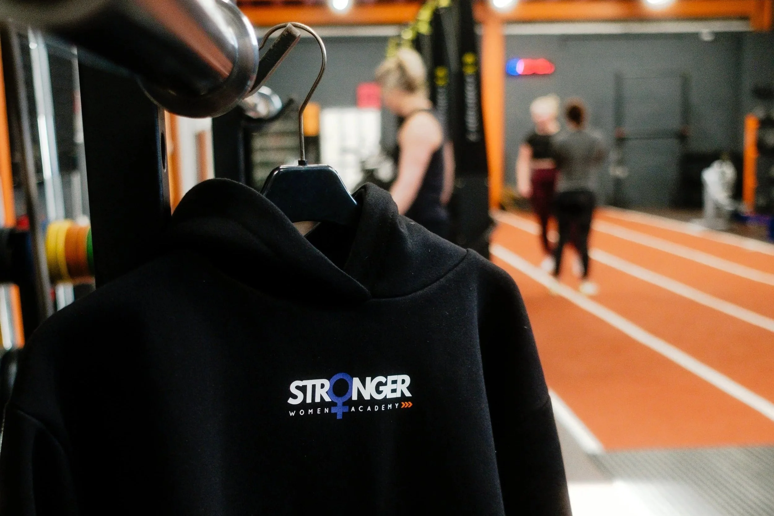 Black hoodie on a hanger with the words 'STRONGER WOMEN'S ACADEMY' printed on it, hanging in a gym with people working out in the background.